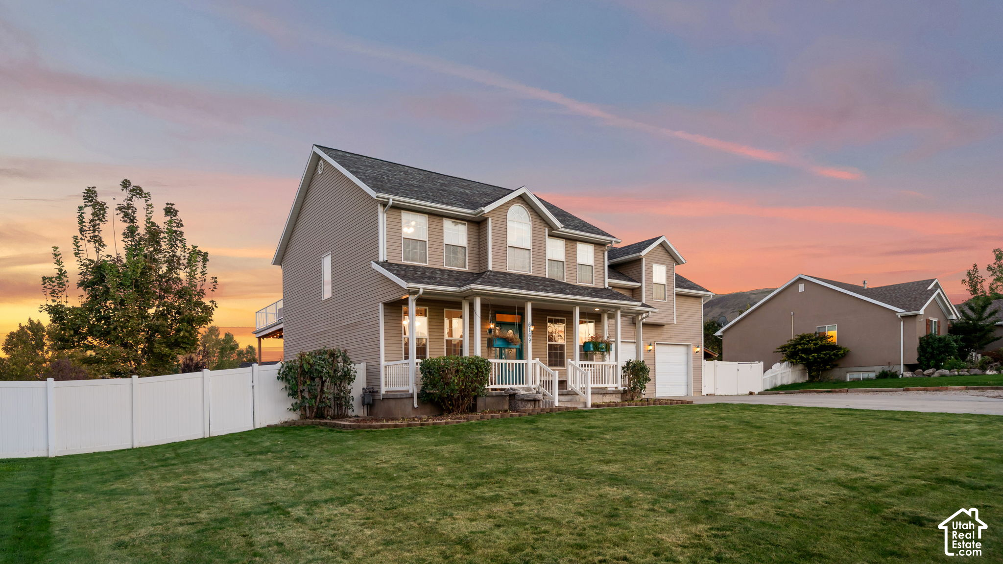 Two-story taupe house with a covered porch and white fence beneath a colorful sunset sky.