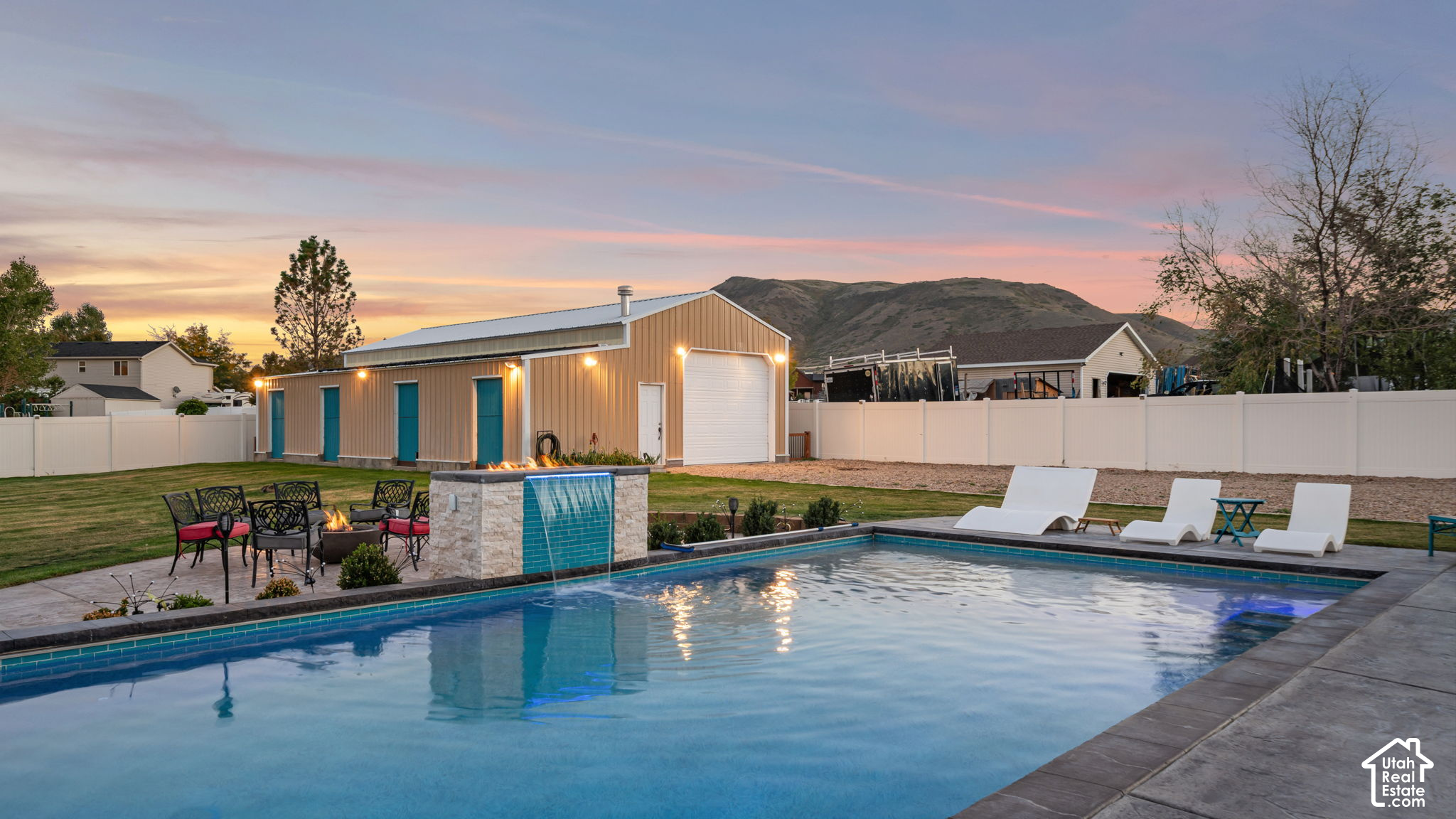 Backyard pool with tiled waterfall, lounge chairs, fire pit, and detached garage at dusk.