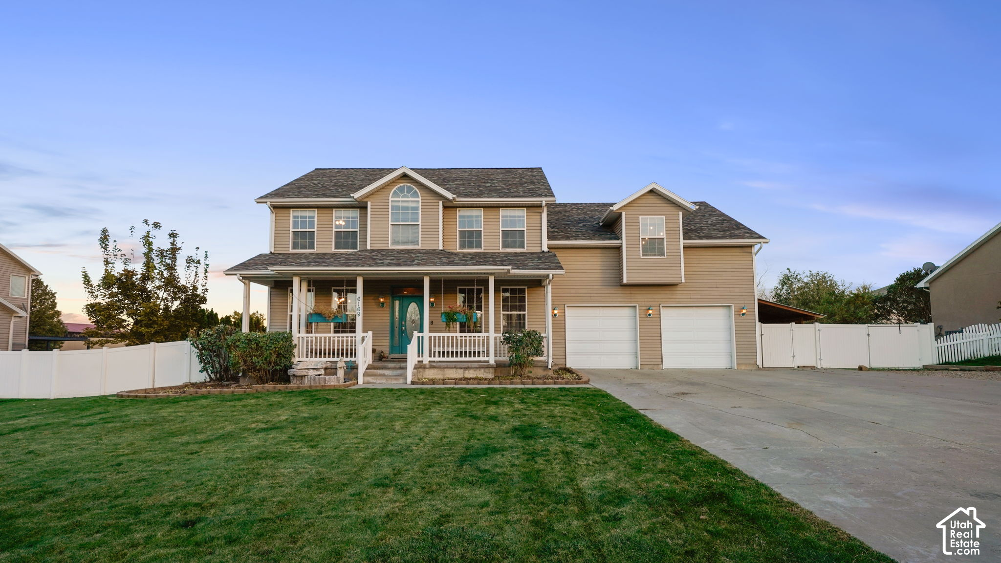 Two-story tan house with a covered porch, teal door, two-car garage, and white fence.