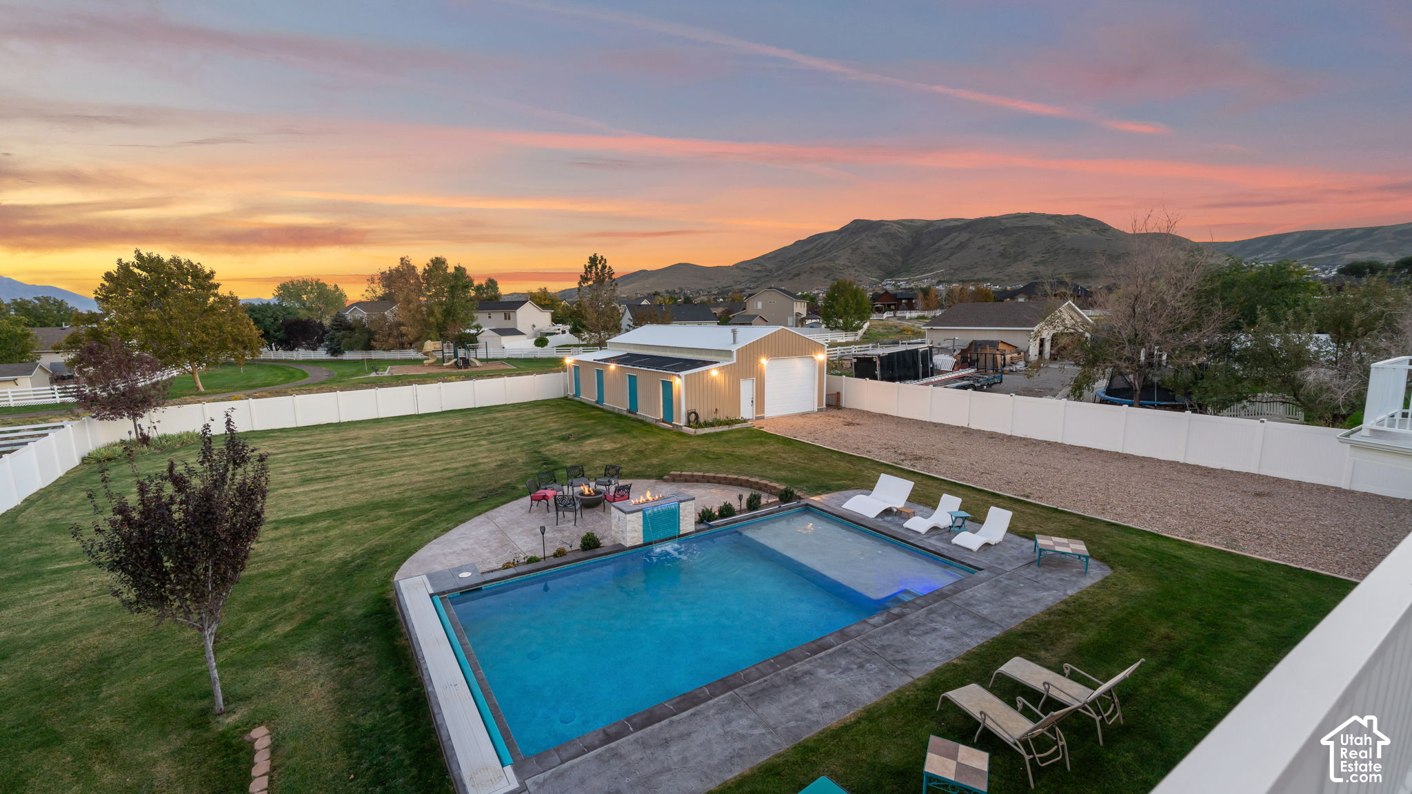 Aerial view of a fenced backyard with a pool, patio, detached outbuilding, and mountains at sunset.