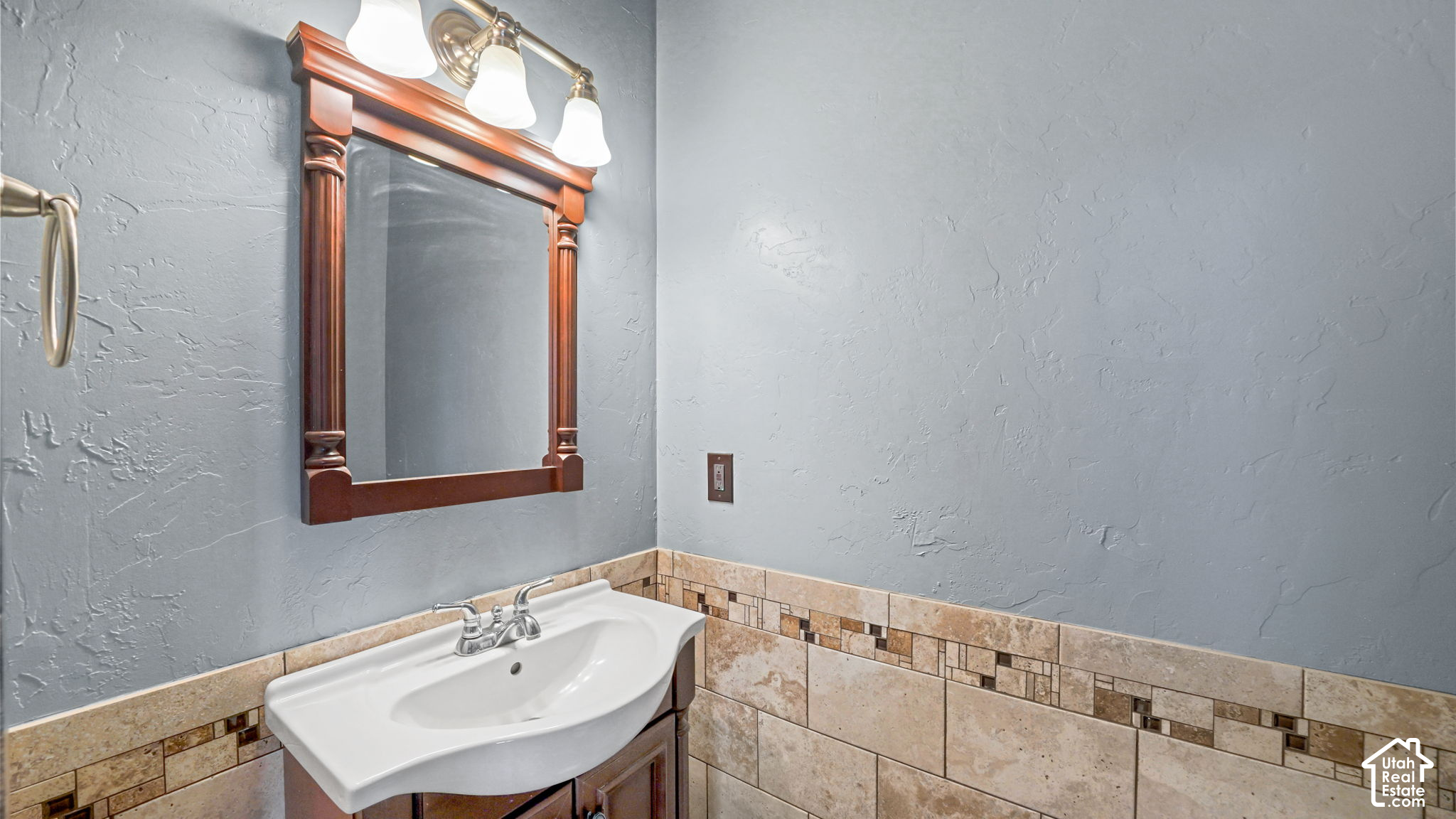 Bathroom vanity with a white sink, framed mirror, three-light fixture, and tan tiled walls.