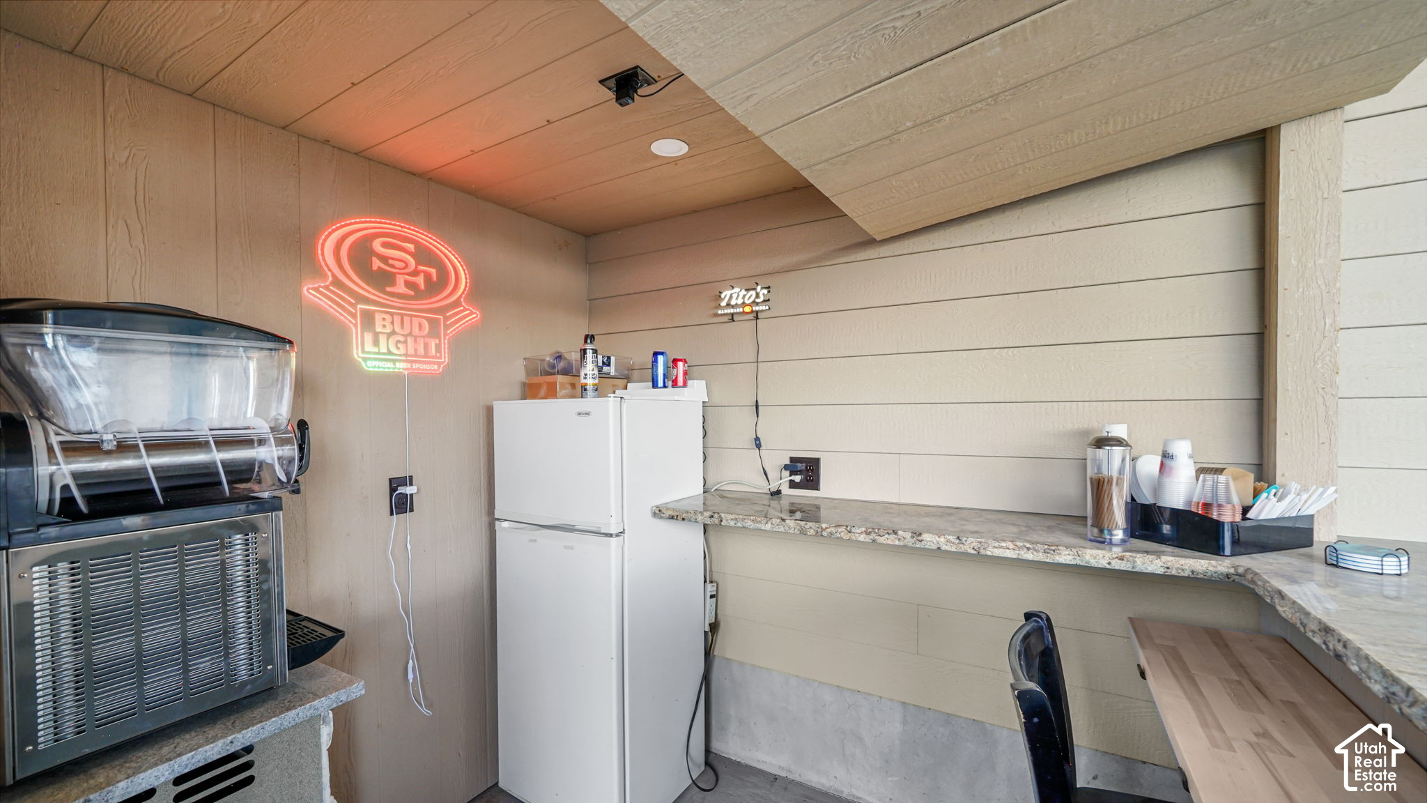 Covered bar area with a granite counter, refrigerator, and commercial frozen drink machine.