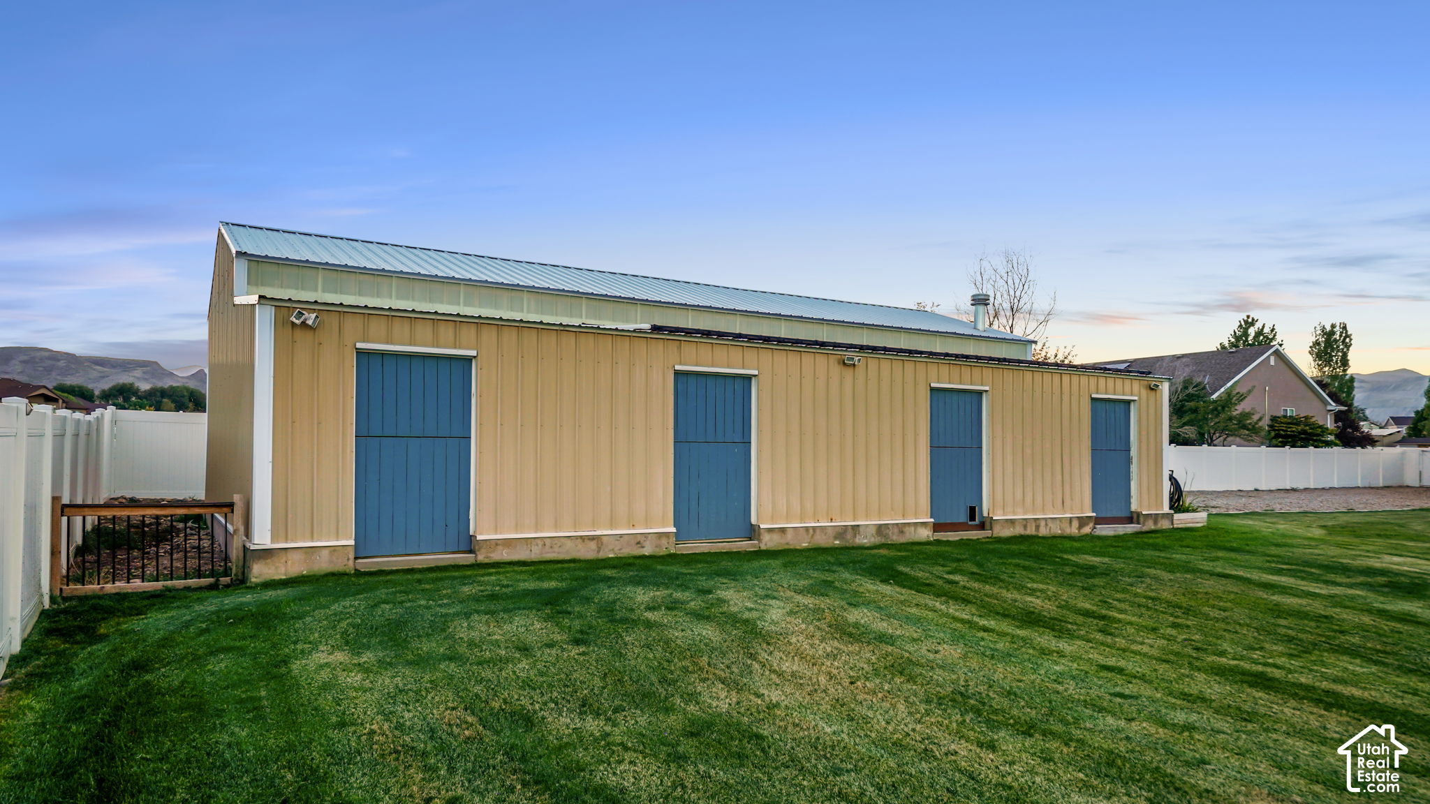 Long tan outbuilding with four blue doors and a pale blue metal roof on a fenced lawn.