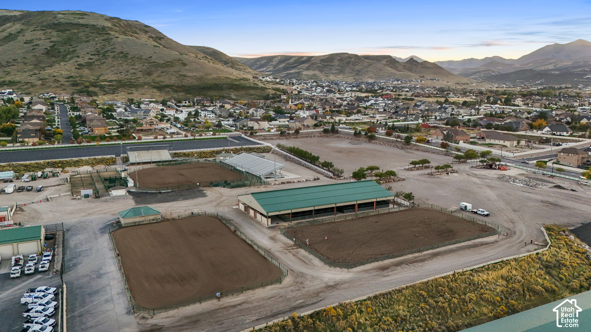 Aerial view of an equestrian facility with riding arenas, green-roofed buildings, homes, and surrounding mountains.
