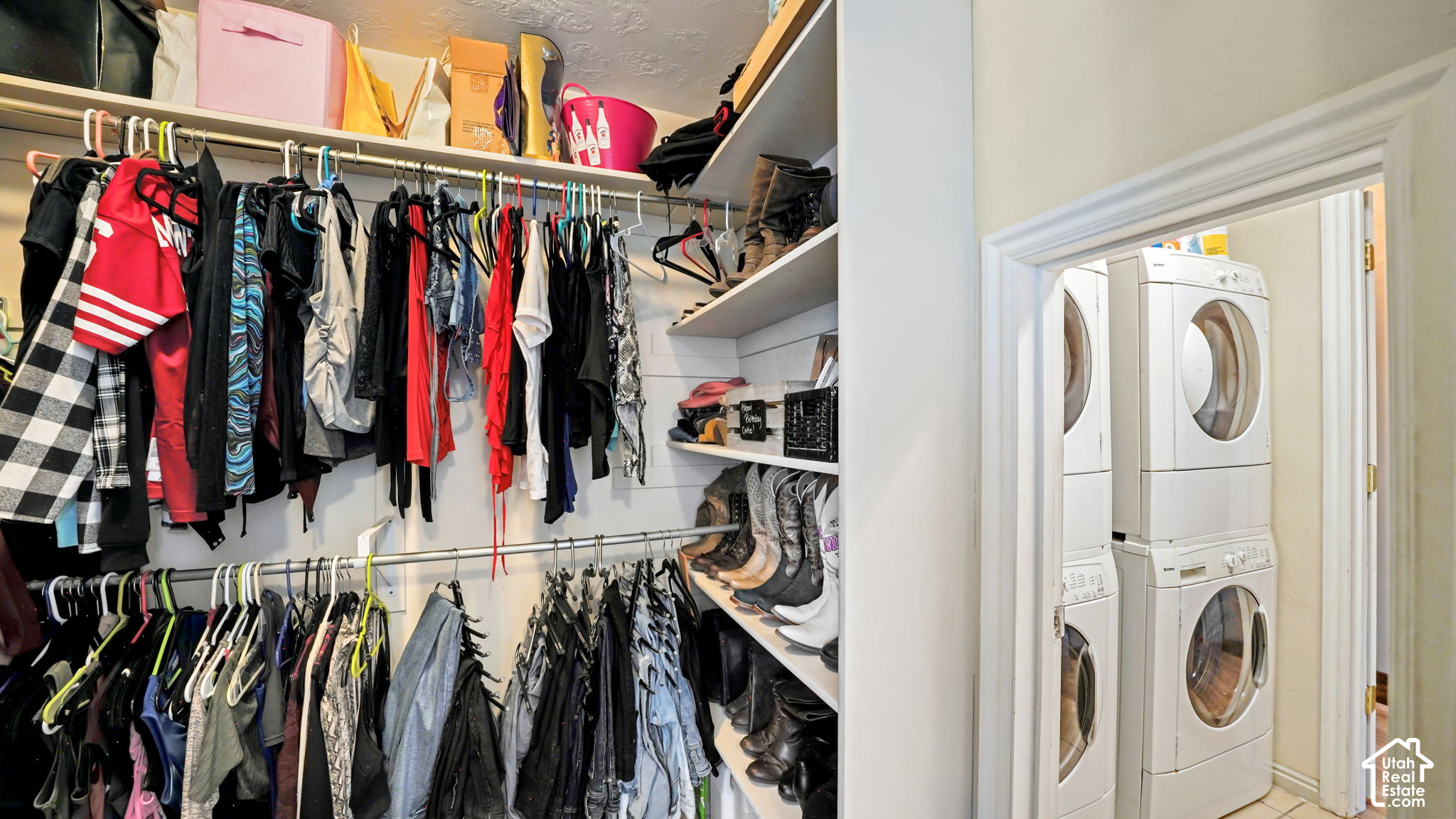 Walk-in closet with two rows of hanging clothes and shoe shelves beside a laundry room with stacked appliances.
