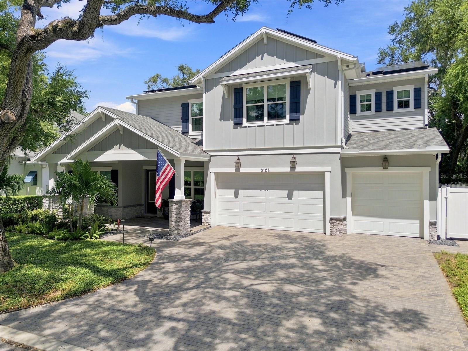 Two-story gray home with solar panels, multiple garages, an American flag, and a broad paver driveway.