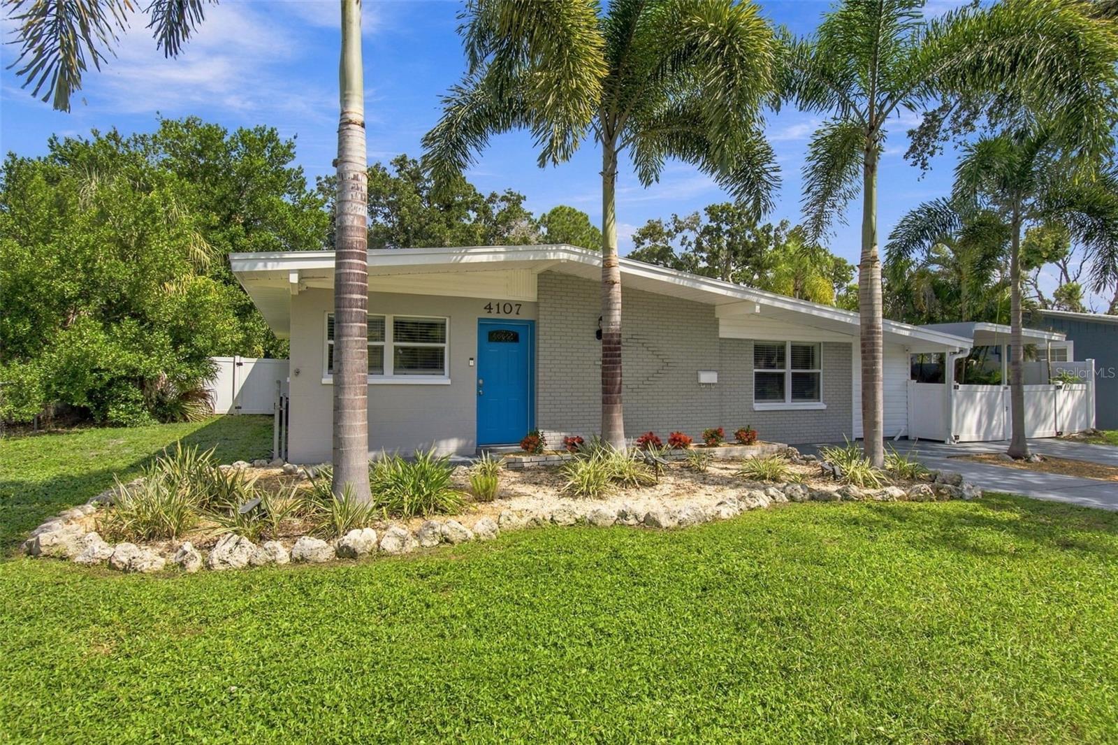 Single-story gray brick house with a bright blue door, palm trees, and a rock-edged garden.