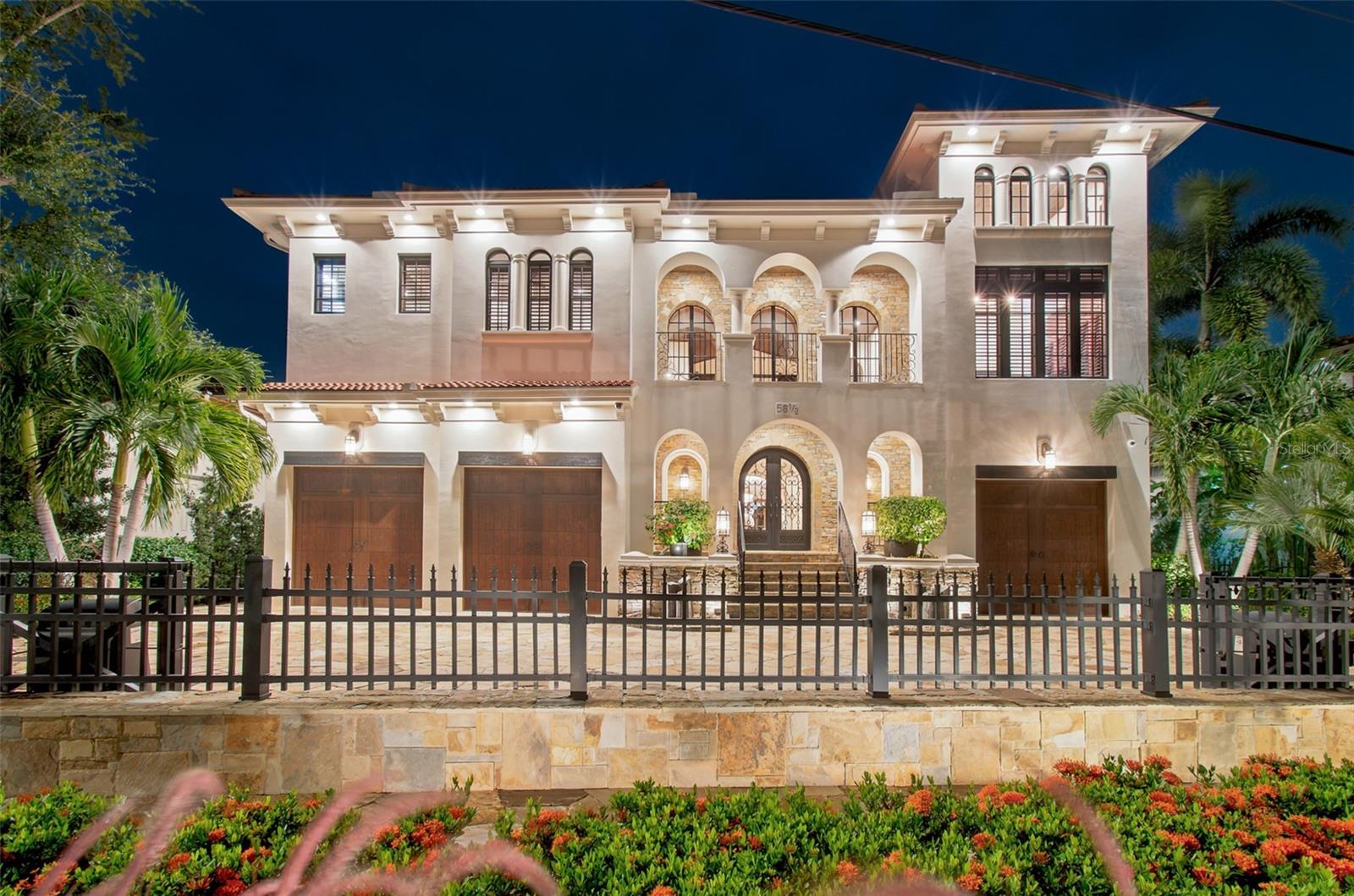 Illuminated two-story home with arched windows, stone accents, three garage doors, and a gated front yard.