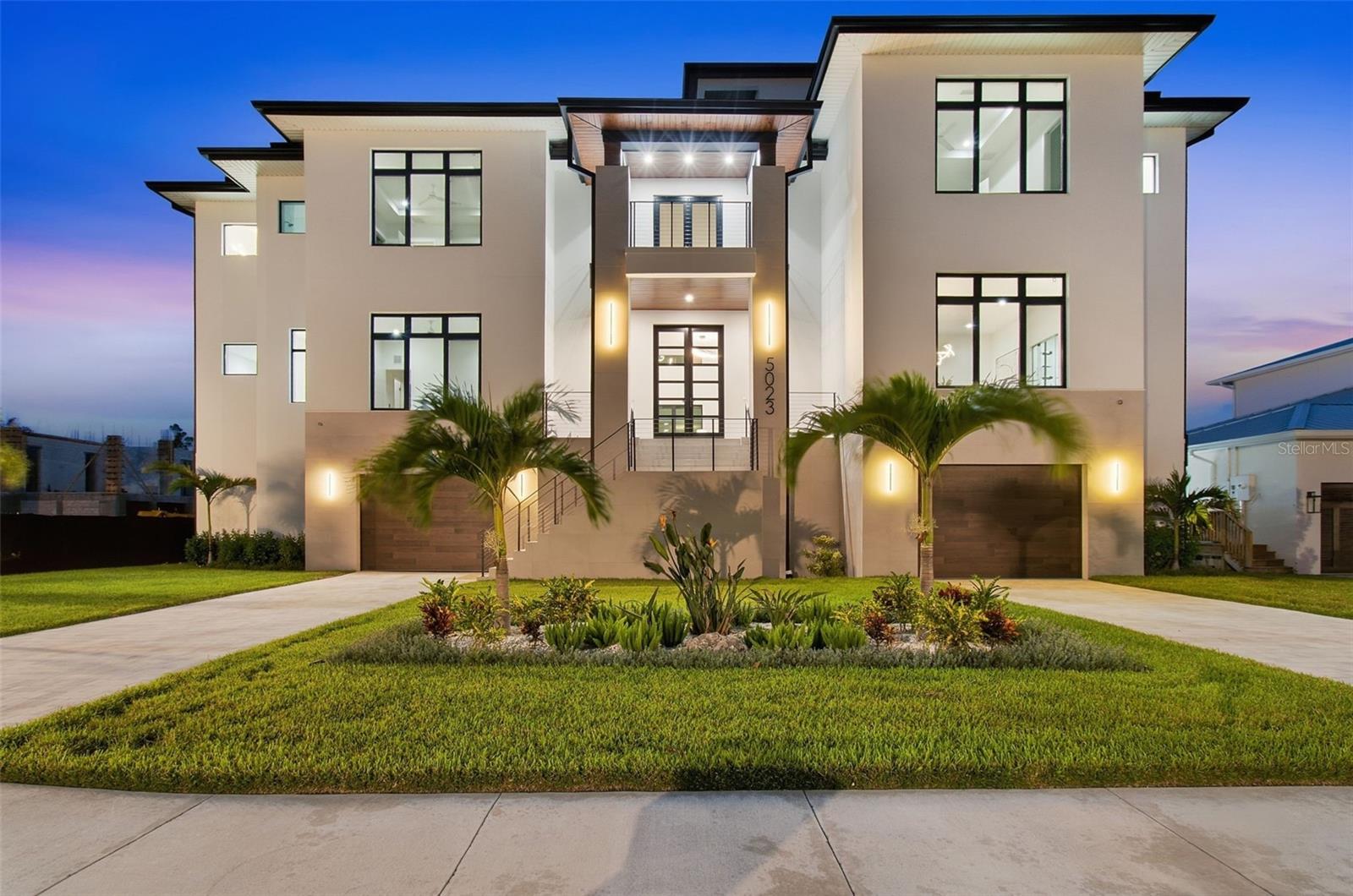 Modern two-story home at dusk with a central entrance, two garages, palm trees, and landscaped lawn.