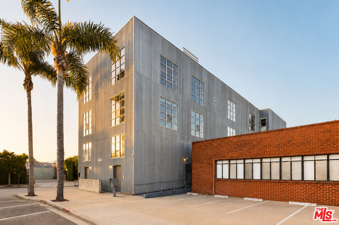 Modern gray multi-story building beside a red-brick structure, with palm trees and parking spaces.