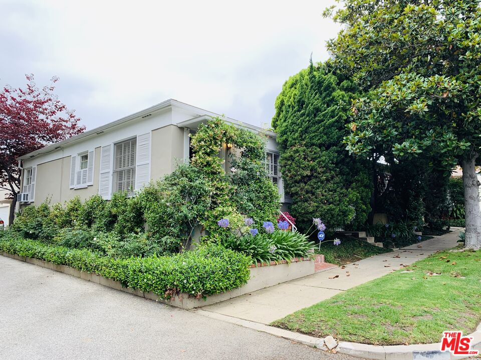White house with shuttered windows, flowering front garden, mature trees, and sidewalk.