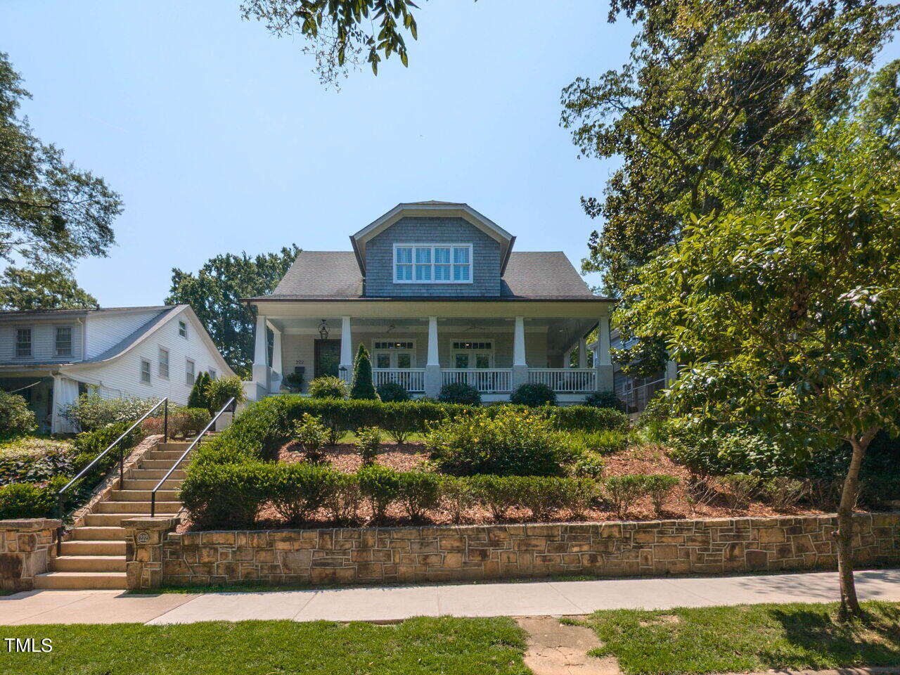 Gray-shingled home with a broad covered porch, landscaped front yard, and stone retaining wall.