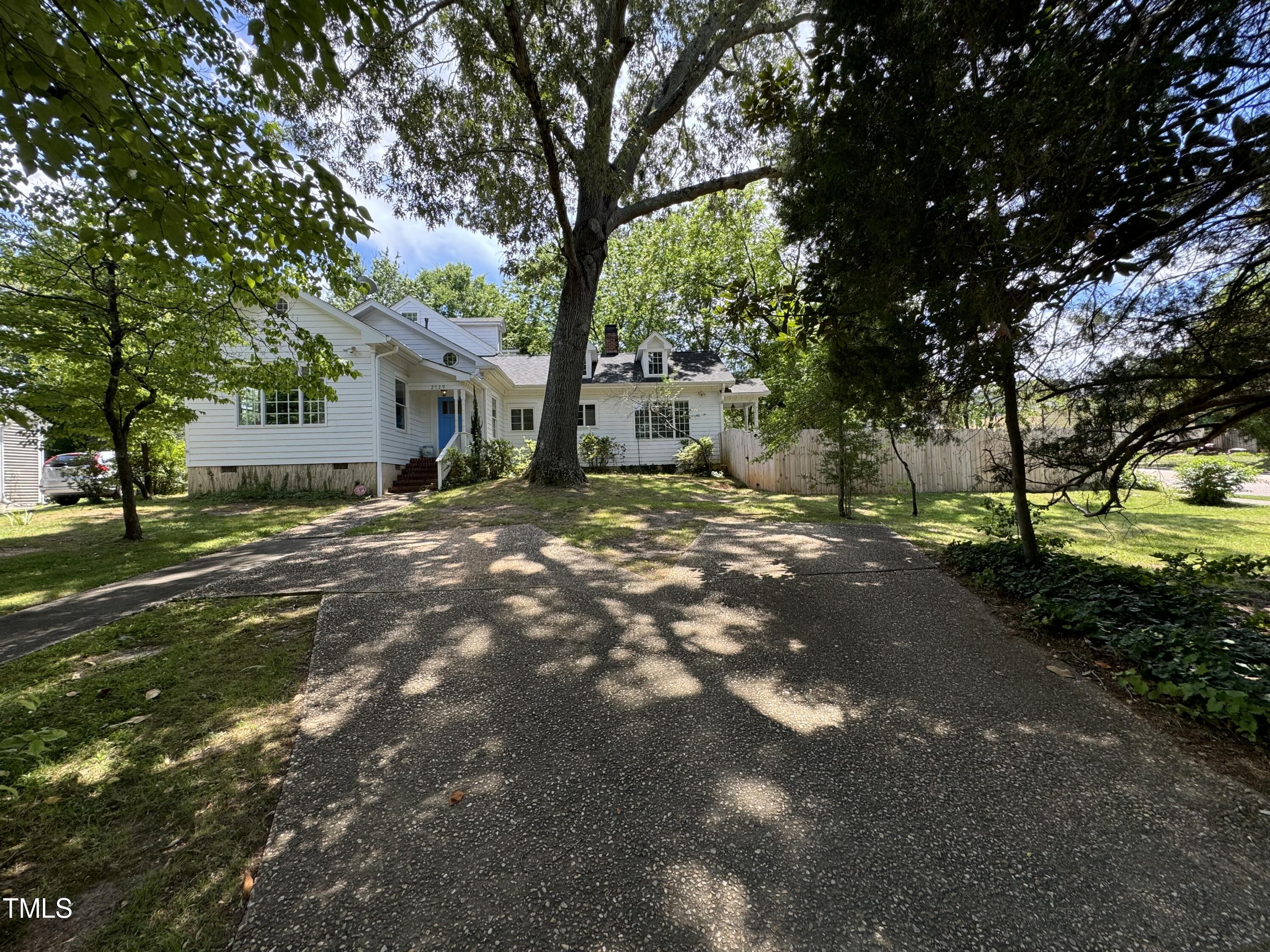 White-sided house with a blue front door behind a wide, tree-shaded driveway.