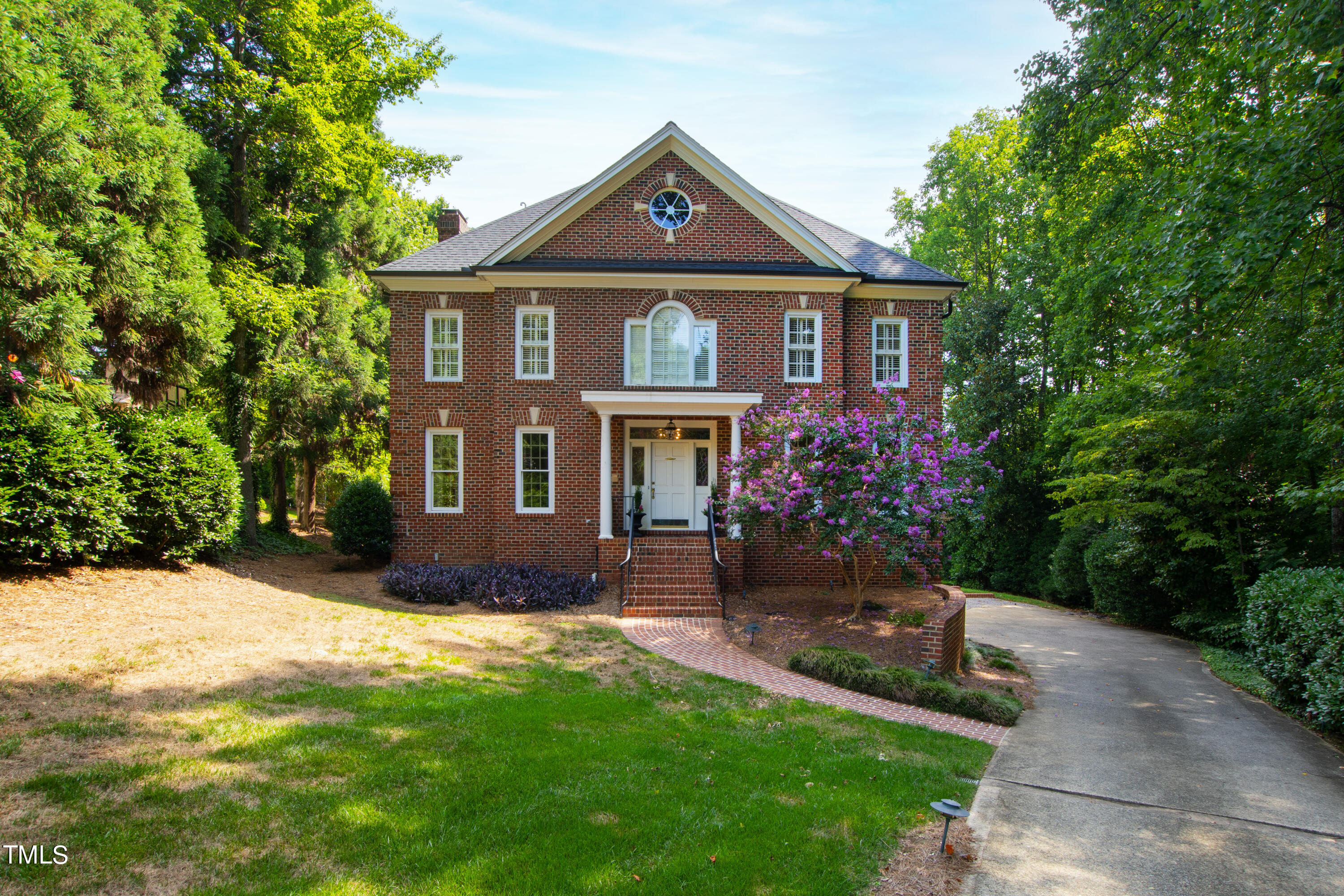 Two-story red brick house with a circular gable window, purple flowering shrubs, and a curving driveway.