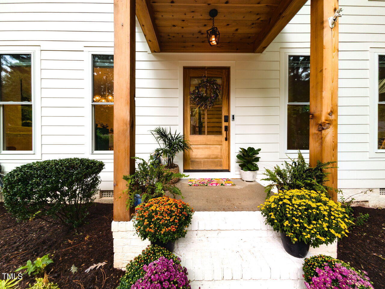 White-sided home with a covered wooden porch, glass-paneled front door, and colorful potted flowers.