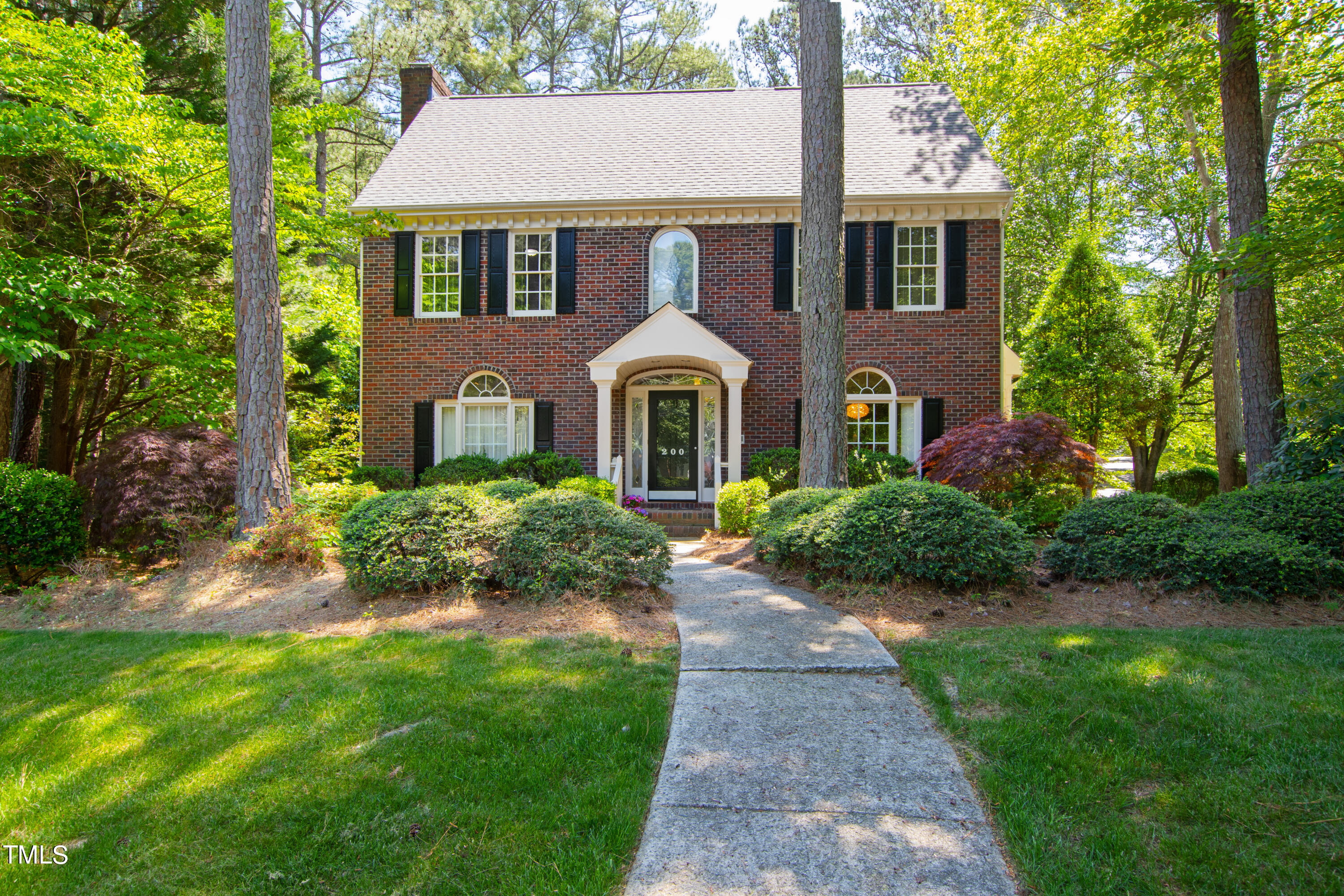 Two-story brick house with black shutters, framed by mature trees and reached by a concrete walkway.