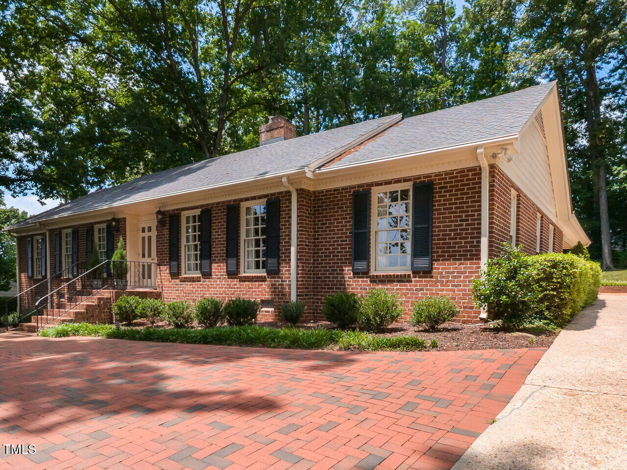 Single-story brick house with black shutters and a broad brick-paver driveway beneath mature trees.