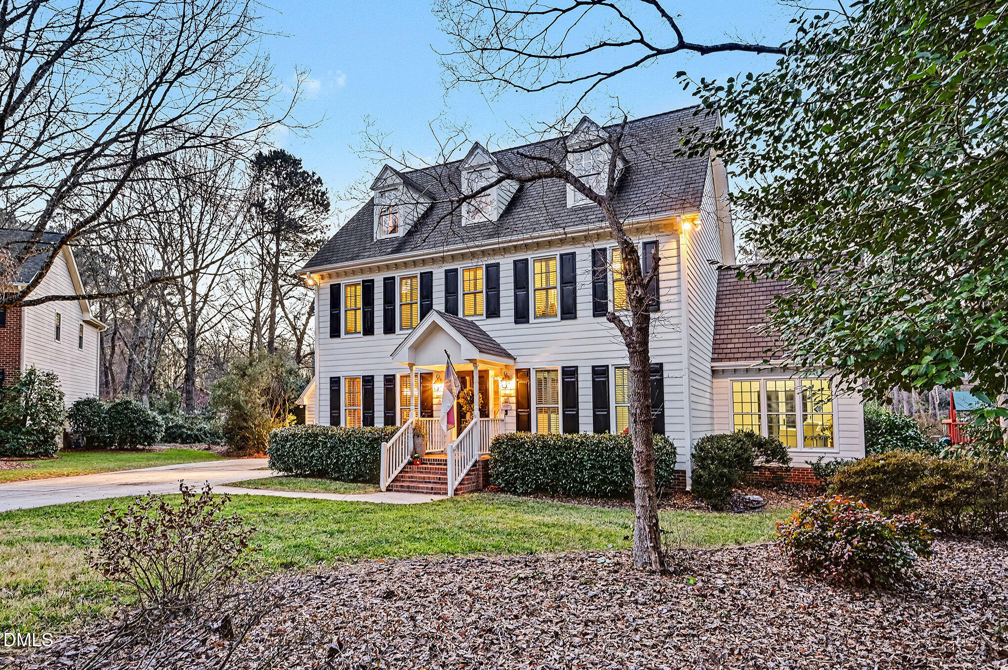 White two-story house with dormers, black shutters, and warmly lit front porch at dusk.