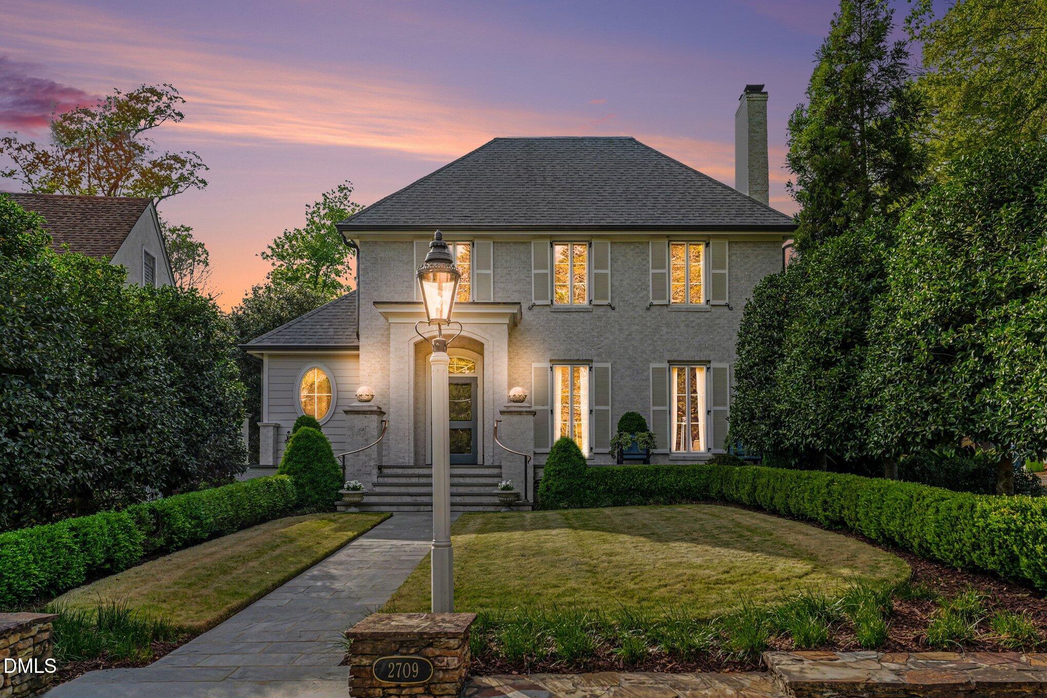 Two-story gray brick home at dusk, with glowing windows, a stone walkway, and a prominent front lamp post.
