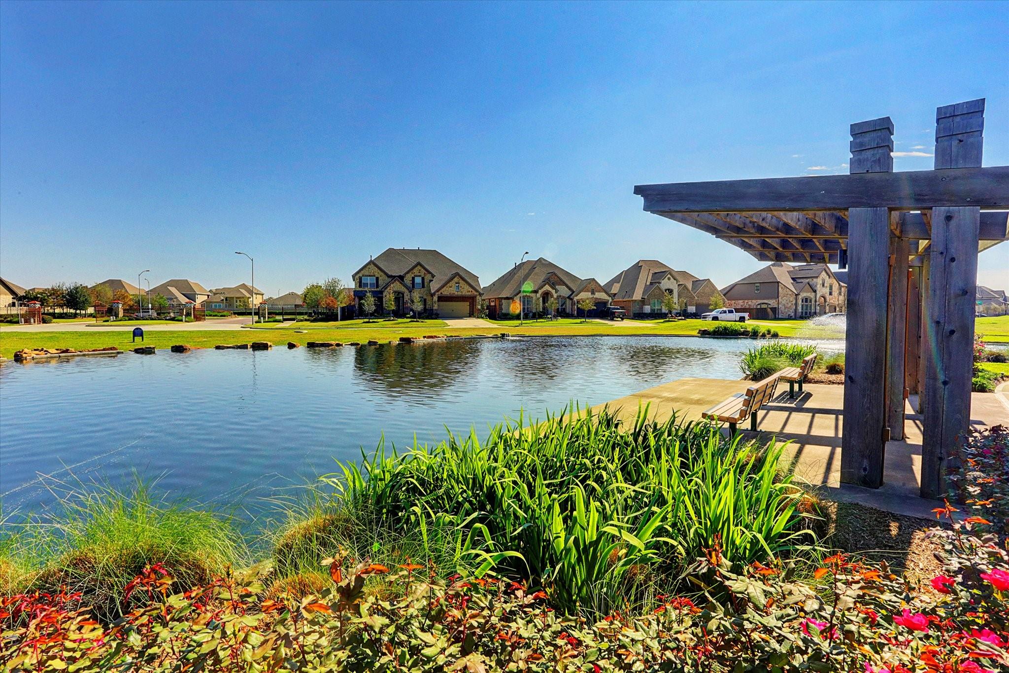 Community pond with a wooden pergola and benches, landscaped banks, and homes across the water.