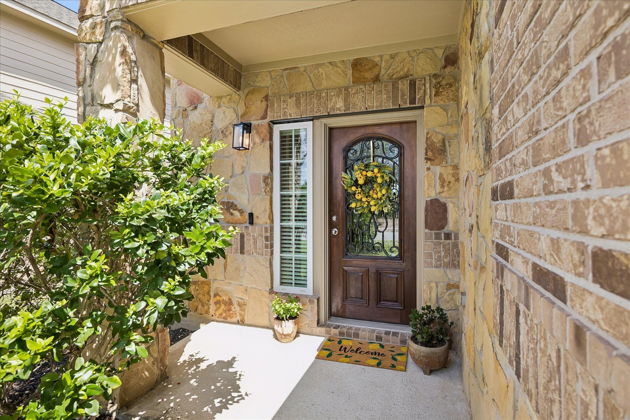 Stone front entry with an ornate wood door, yellow floral wreath, potted plants, and leafy shrubs.