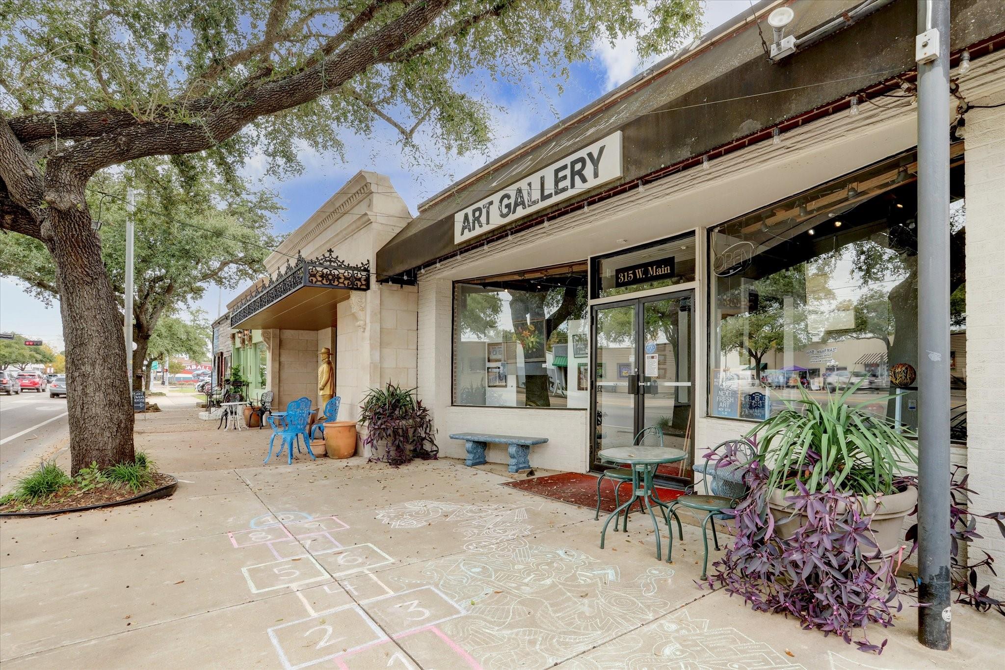 Art gallery storefront with sidewalk seating, potted plants, and chalk drawings beneath a large shade tree.