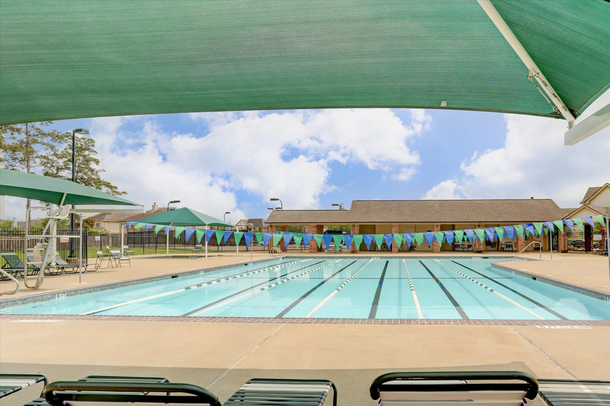 Community lap pool with lane ropes, colorful pennant banners, and green shade canopies.