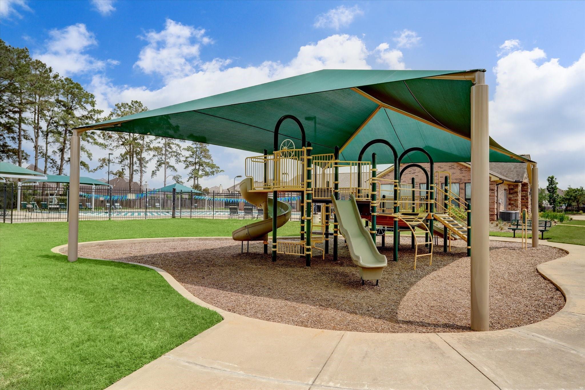 Neighborhood playground with shaded climbing structure and slides, with a fenced pool in the background.