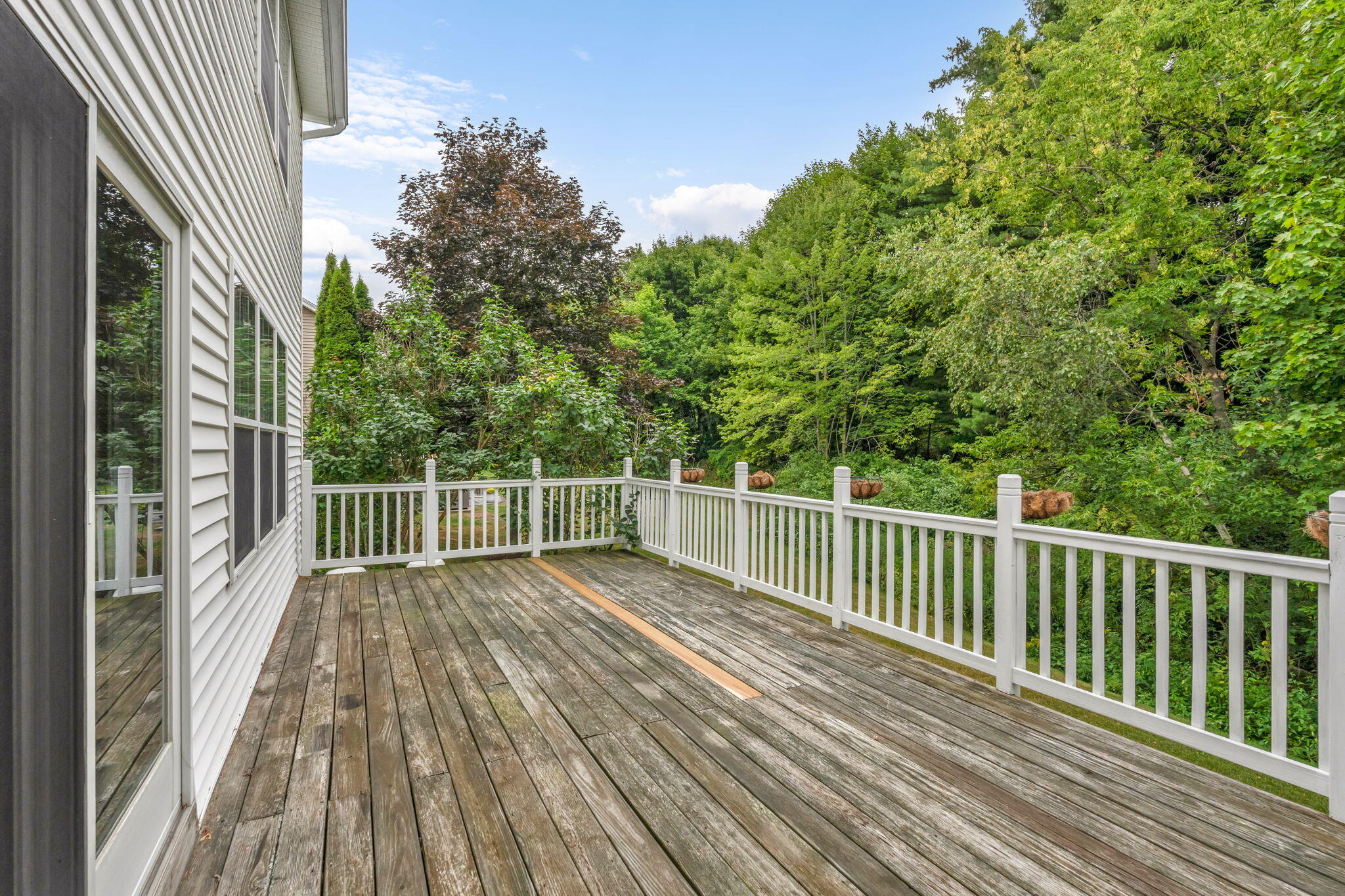 Wood deck with white railings overlooking dense green trees beside a light-colored home.