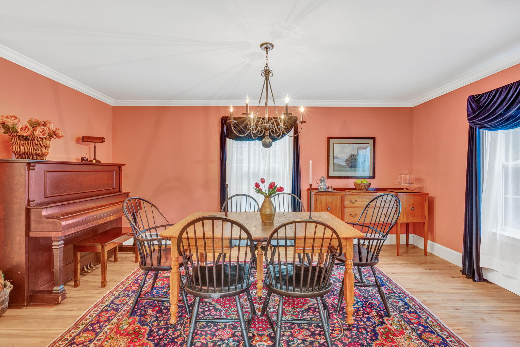 Dining room with a wooden table, colorful rug, upright piano, and chandelier.