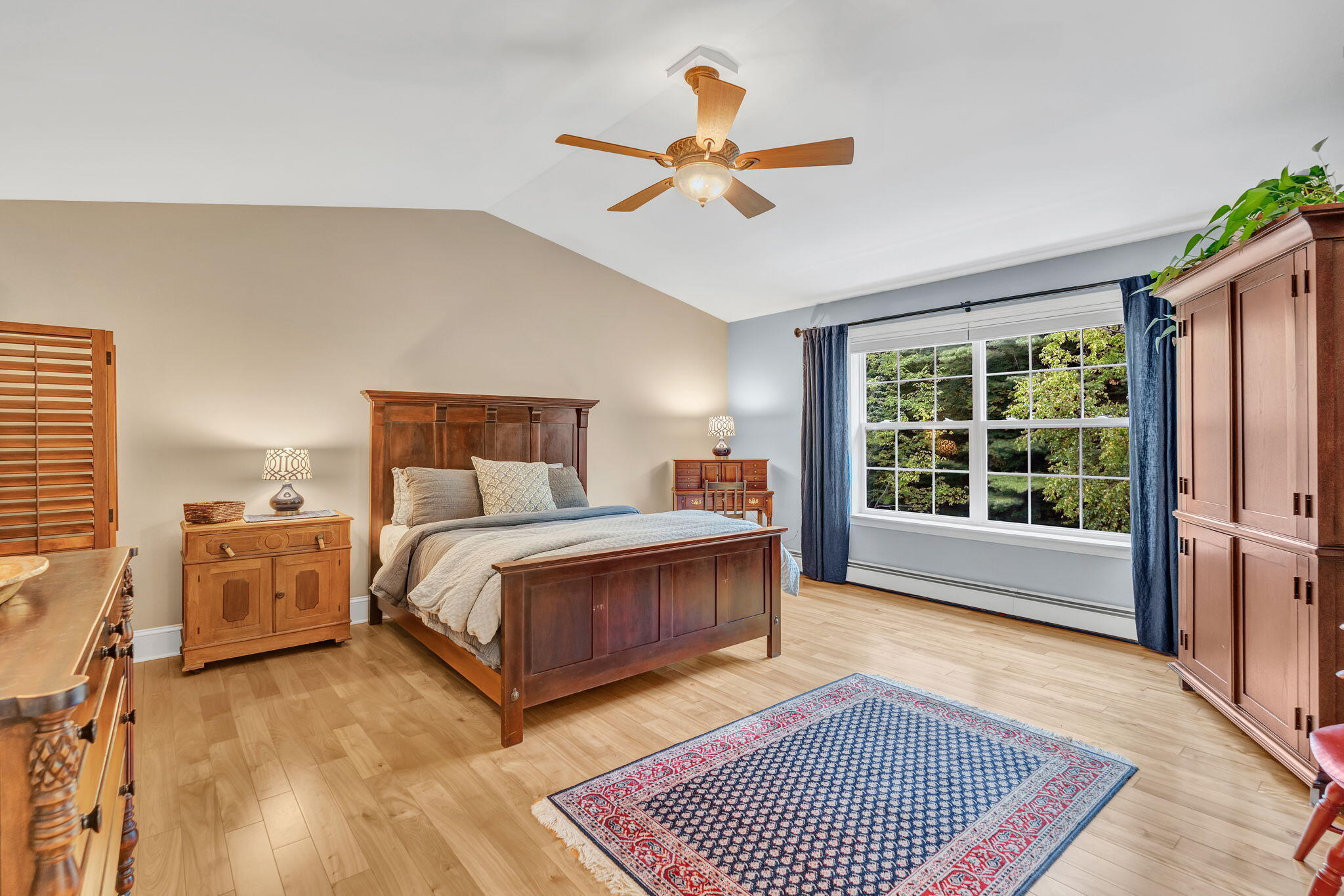 Furnished bedroom with a large wooden bed, patterned rug, and wide window overlooking trees.