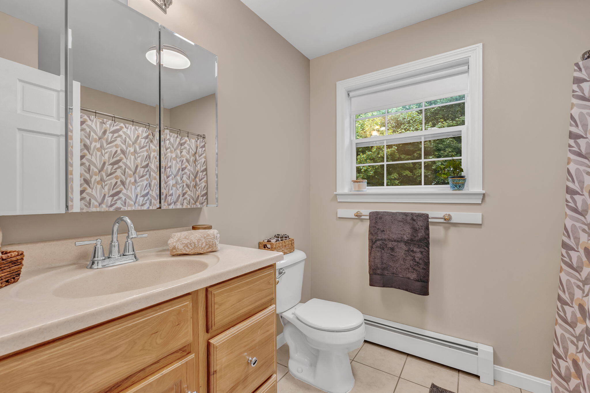 Bathroom with a wood vanity and mirrored cabinet, patterned shower curtain, and window facing leafy greenery.