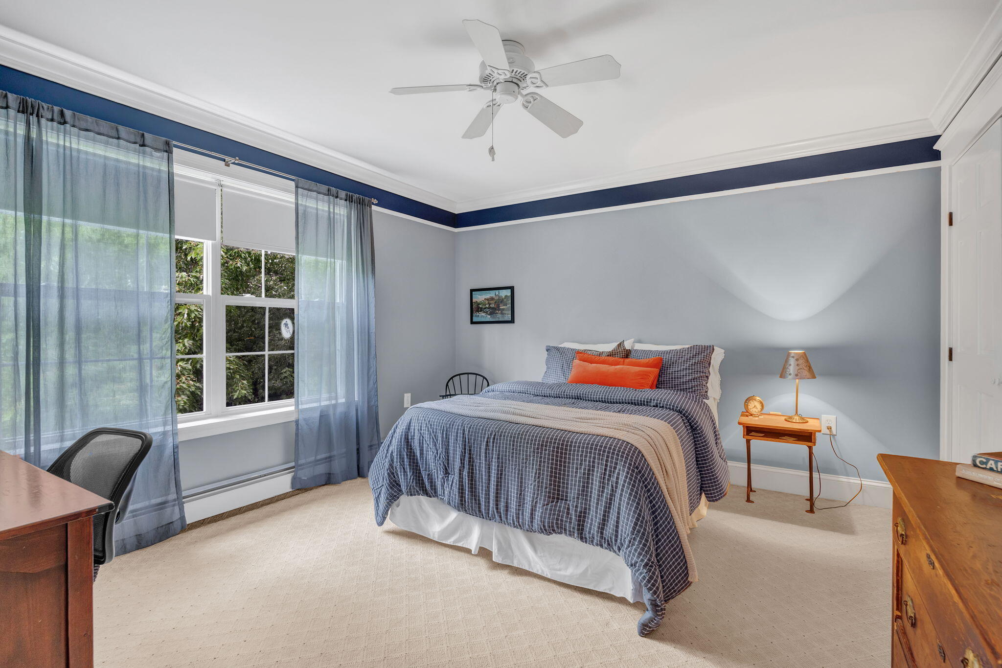 Bedroom with blue checked bedding, orange accent pillow, and large curtained windows.