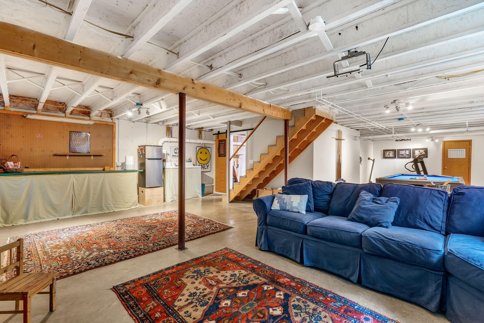 Basement recreation room with a blue sectional, pool table, patterned rugs, and open wooden stairs.