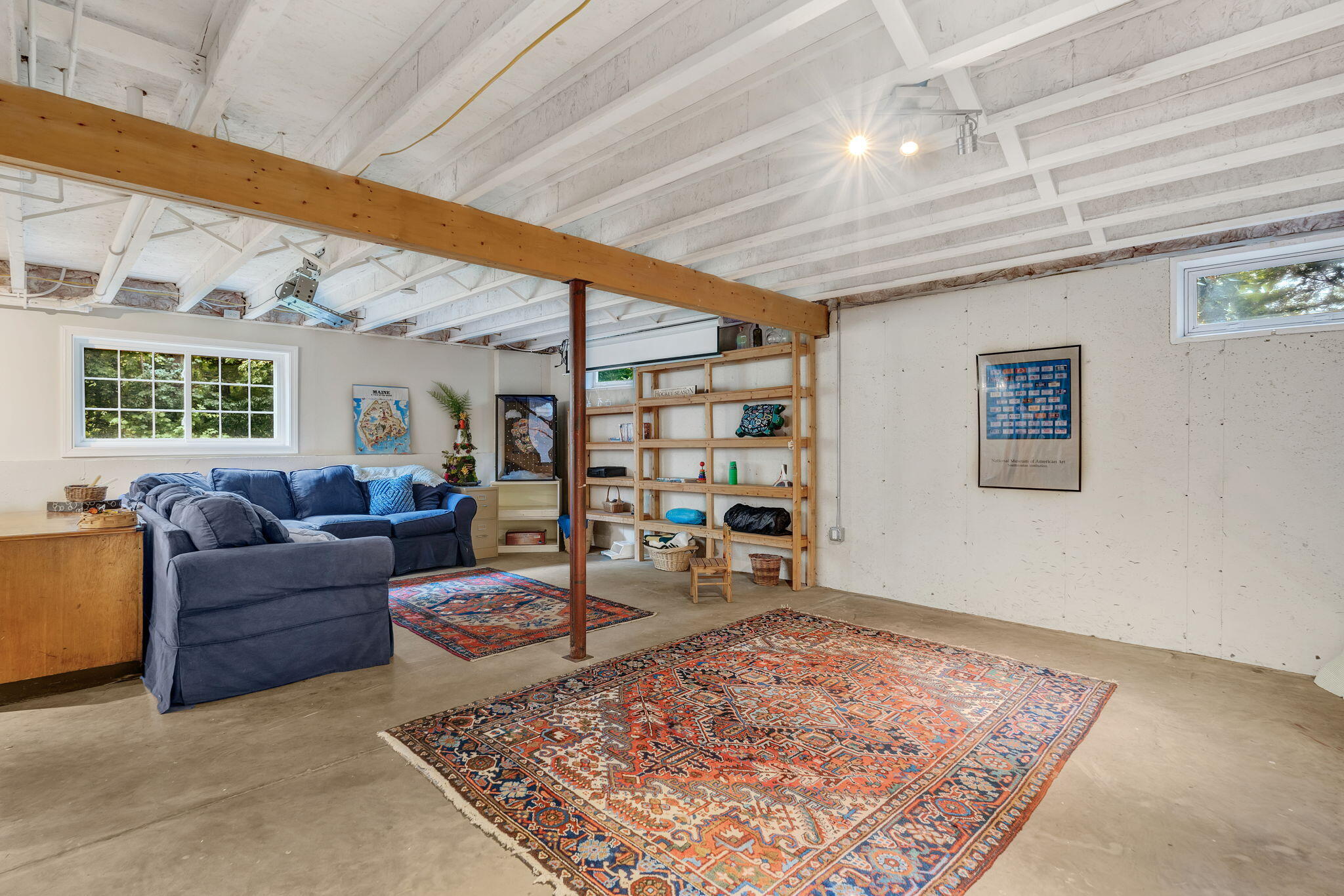 Furnished basement living area with a blue sectional, exposed beams, open shelving, and patterned rugs.