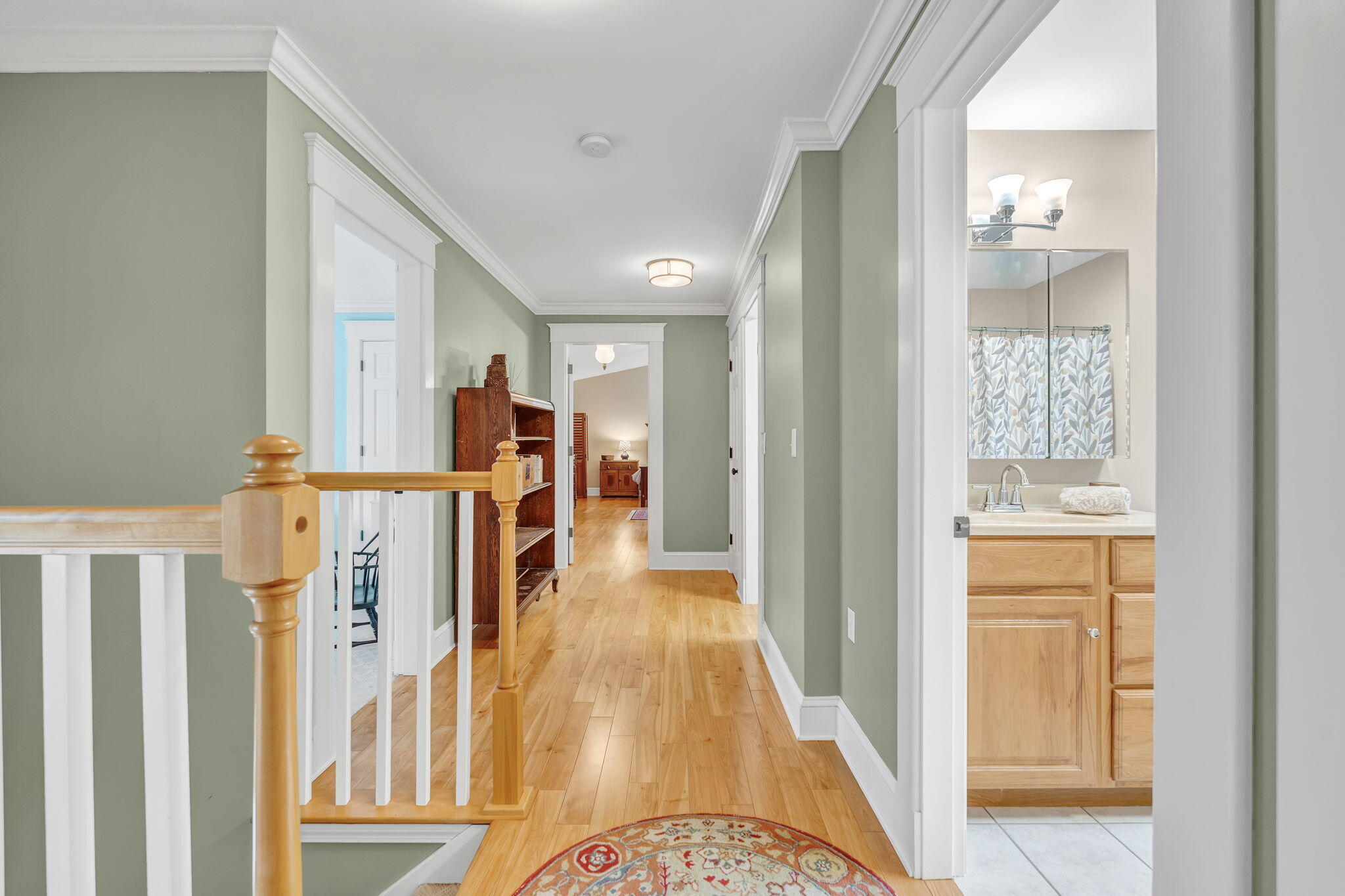 Hallway with sage-green walls and hardwood floors, opening to a bathroom with a light wood vanity.