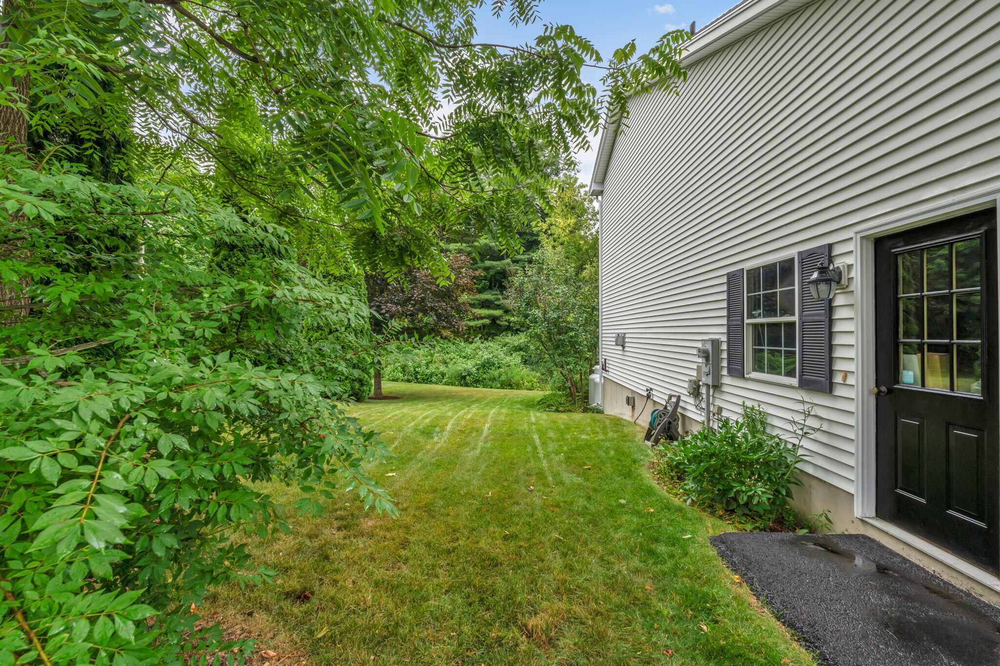 Side entrance of a white-sided home beside a leafy, tree-lined lawn.