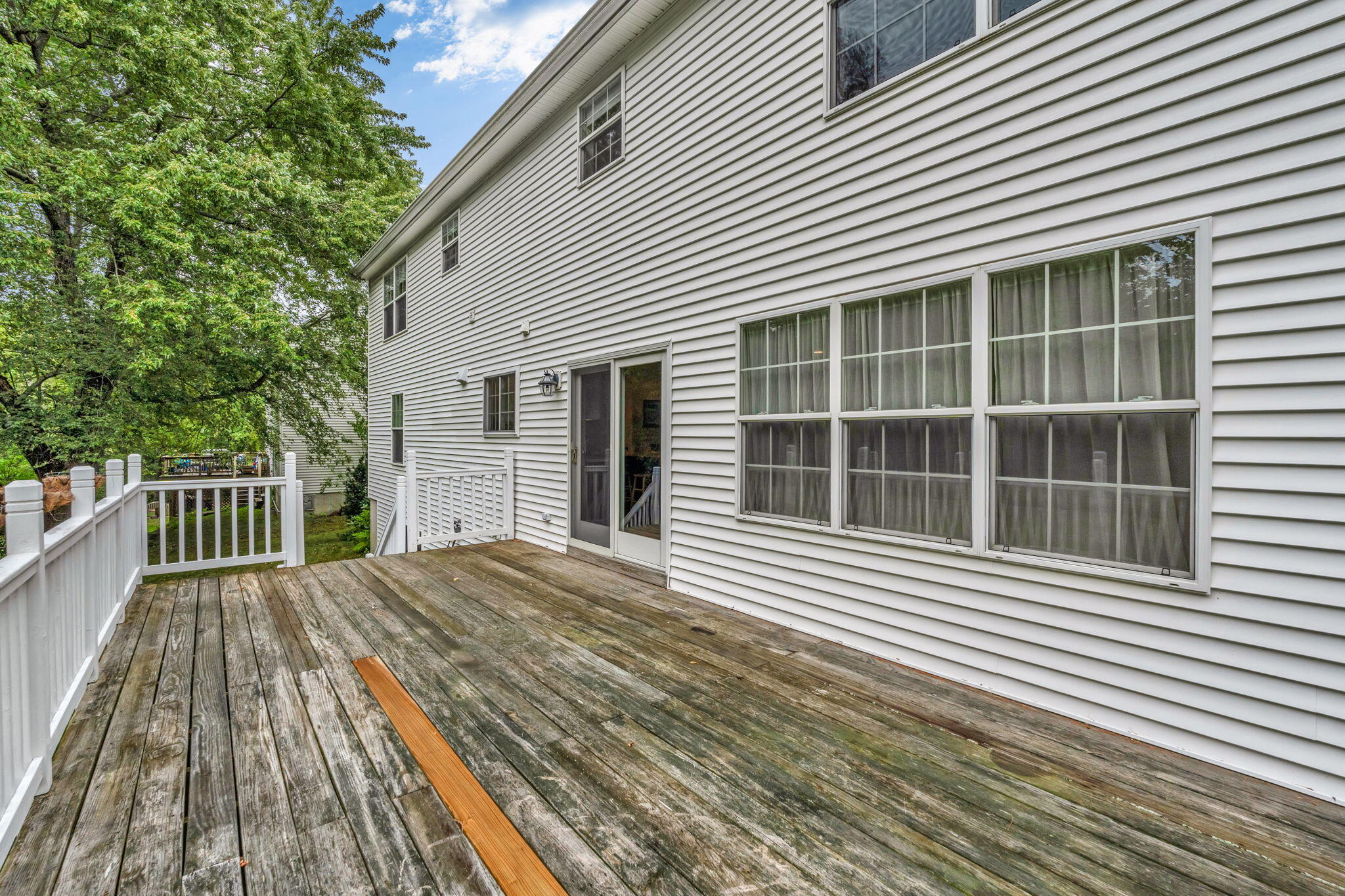 Weathered wooden deck beside a white-sided house, with white railings and leafy trees.
