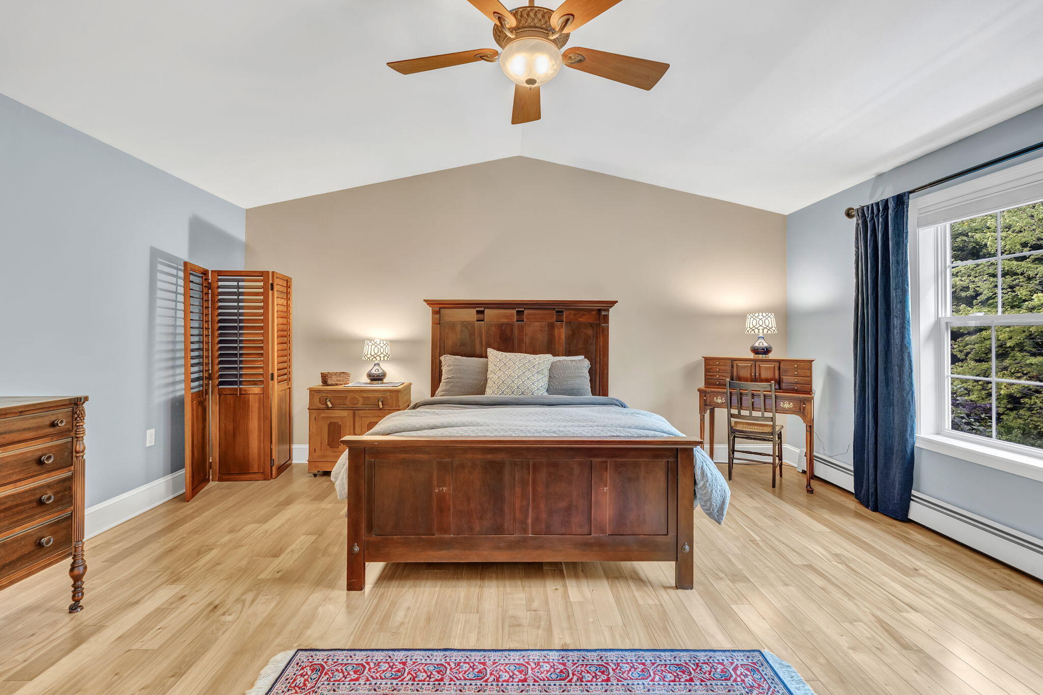 Furnished bedroom with a wooden bed, hardwood floors, ceiling fan, and large curtained window.