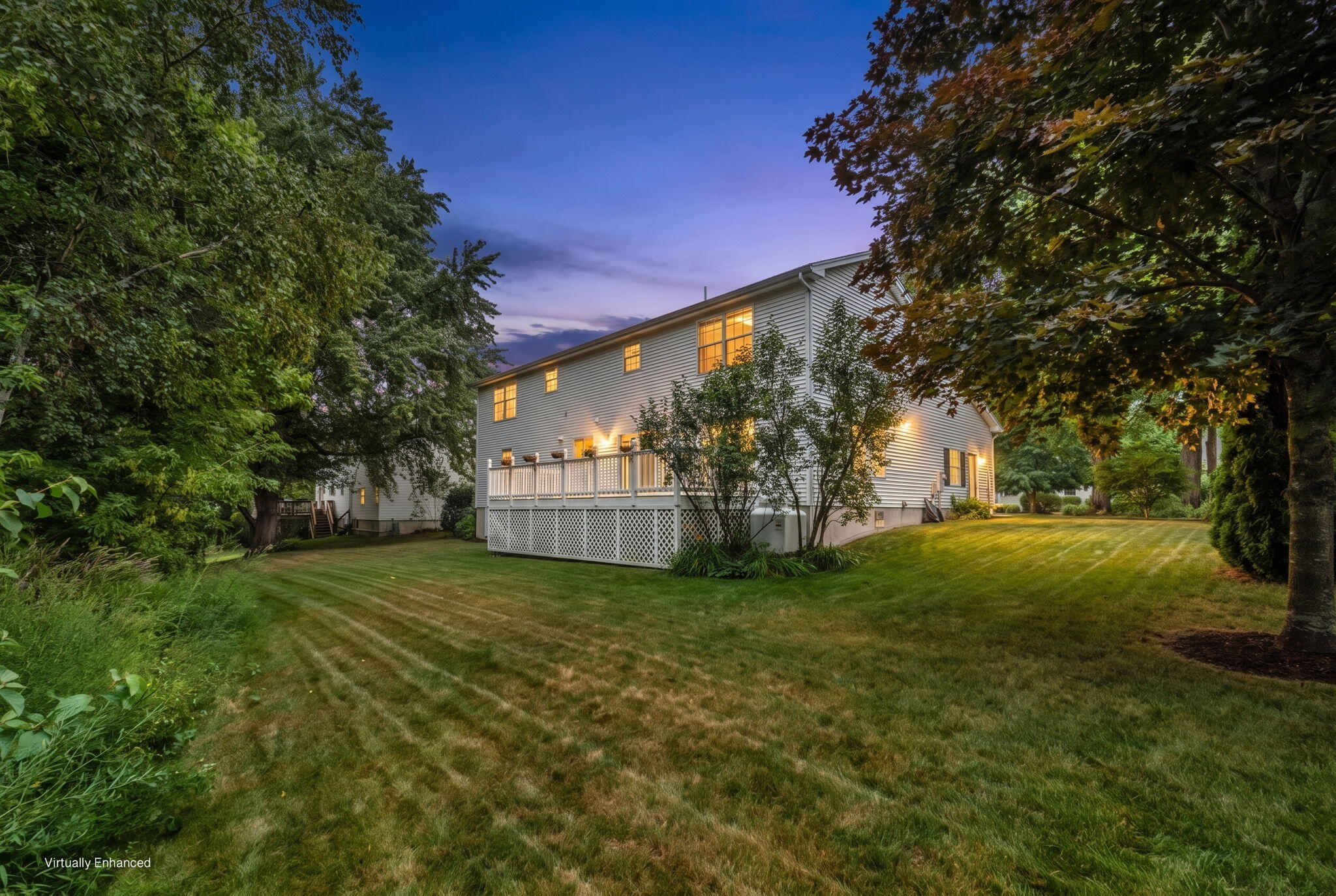 Two-story white house at dusk, with a raised deck and broad tree-lined lawn.