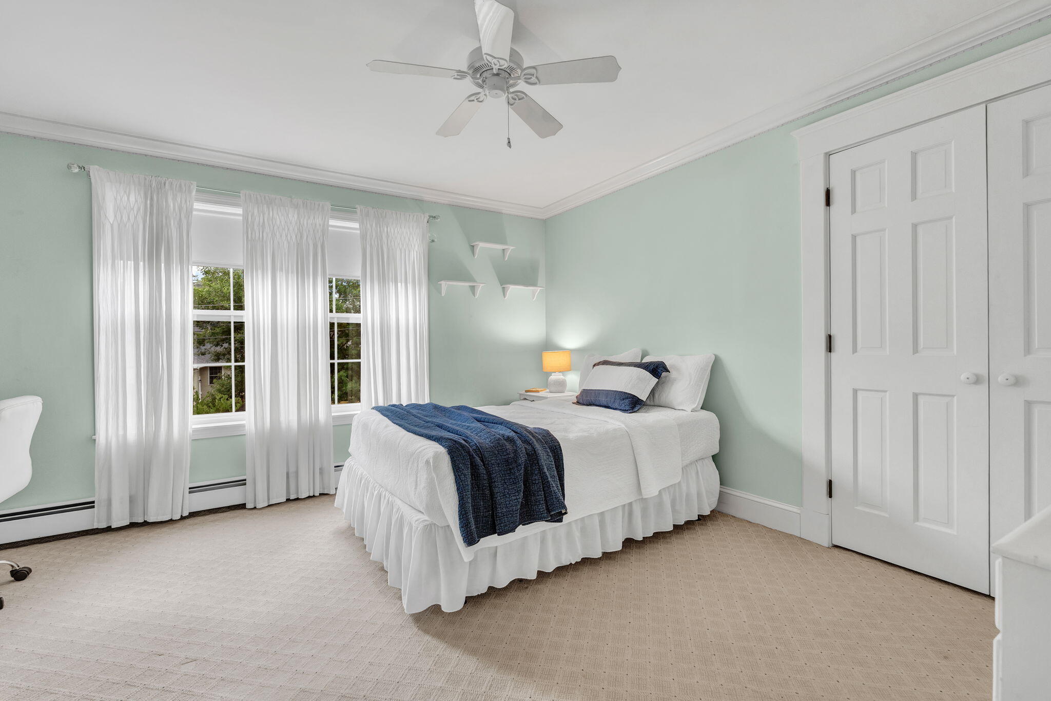 Bedroom with a white bed and navy throw, mint-green walls, and two curtained windows.