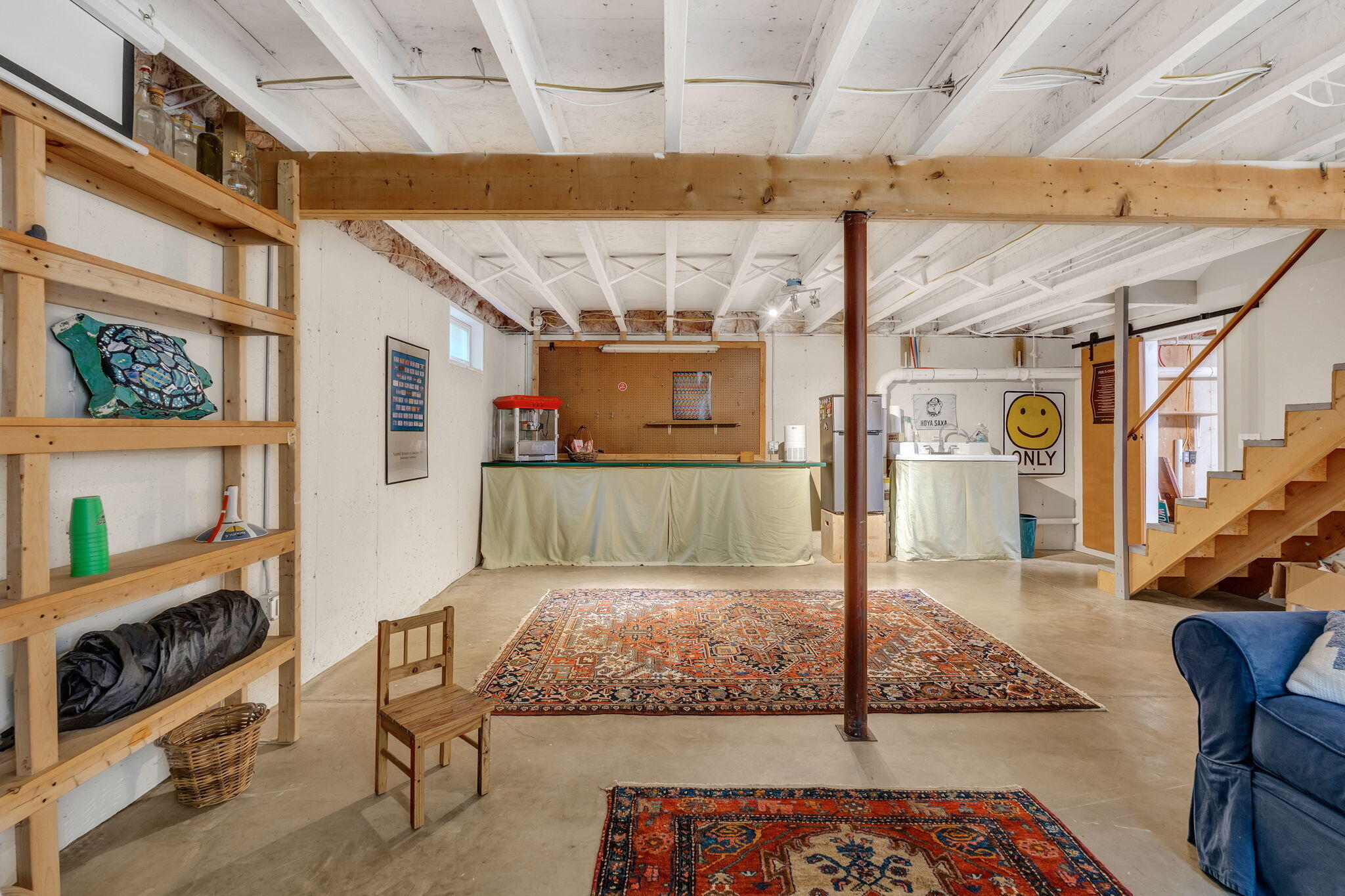 Basement with exposed ceiling joists, built-in shelving, patterned rugs, and stairs.
