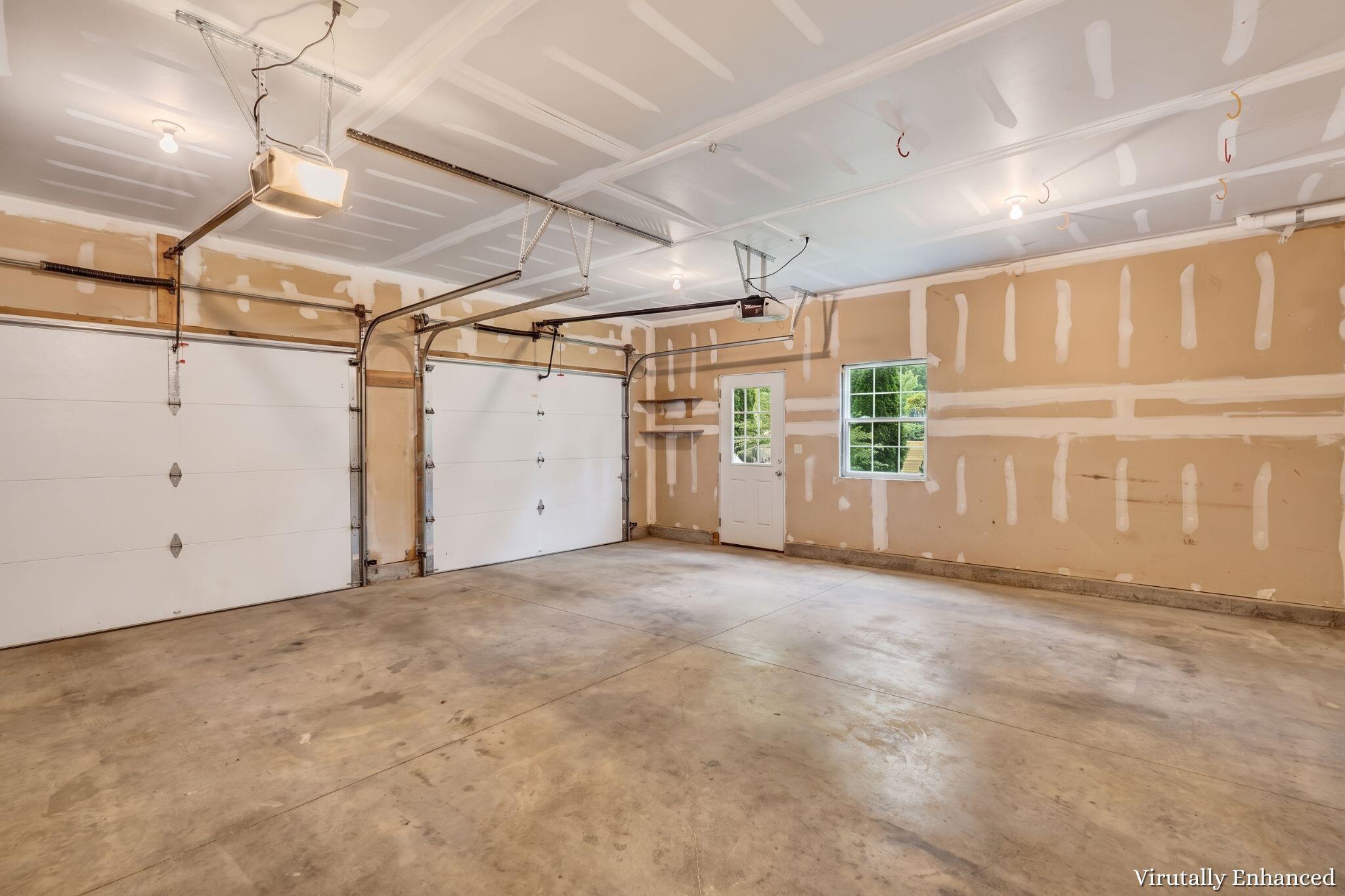 Unfinished two-car garage with white overhead doors, concrete floor, and partially taped drywall.