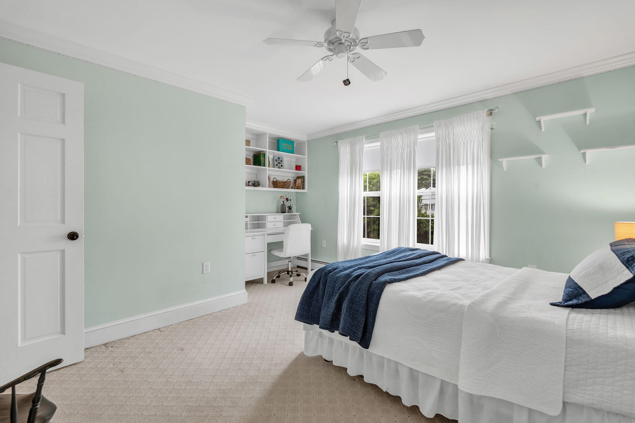 Bedroom with a white bed and navy blanket, pale green walls, and a built-in desk beside curtained windows.