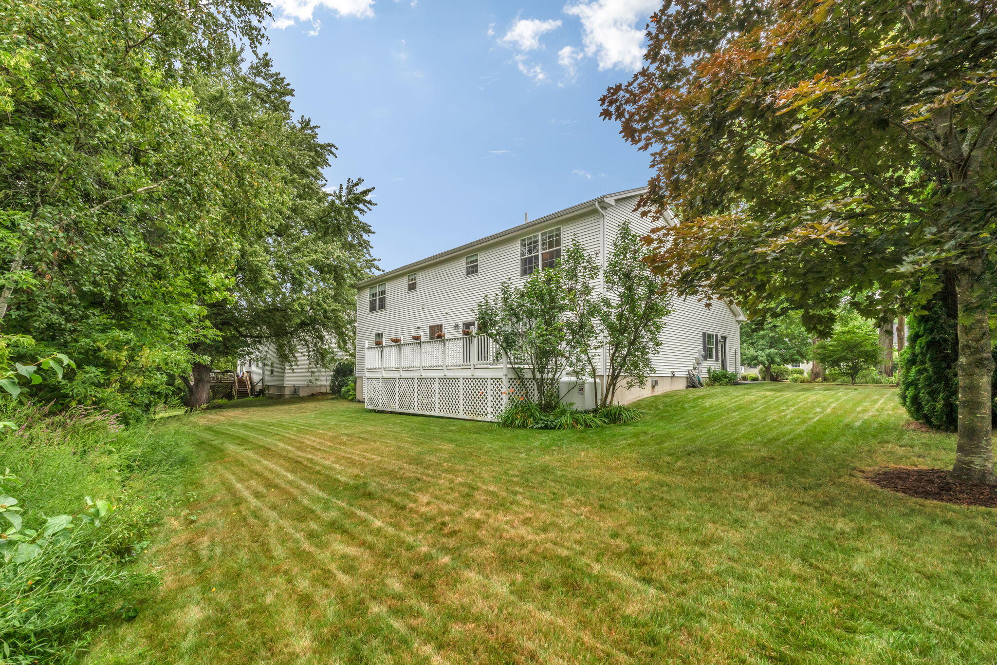 White two-story house with a raised deck behind a broad lawn and mature trees.