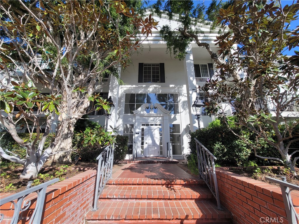 White multi-story building entrance at the top of brick steps, framed by mature trees.