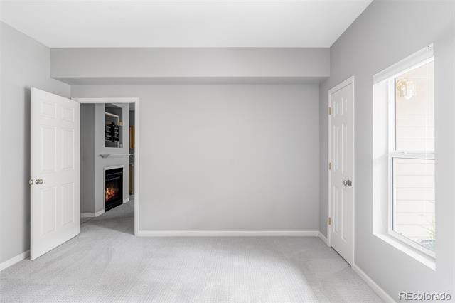 Empty carpeted bedroom with a large window and an open doorway to a room with a fireplace.