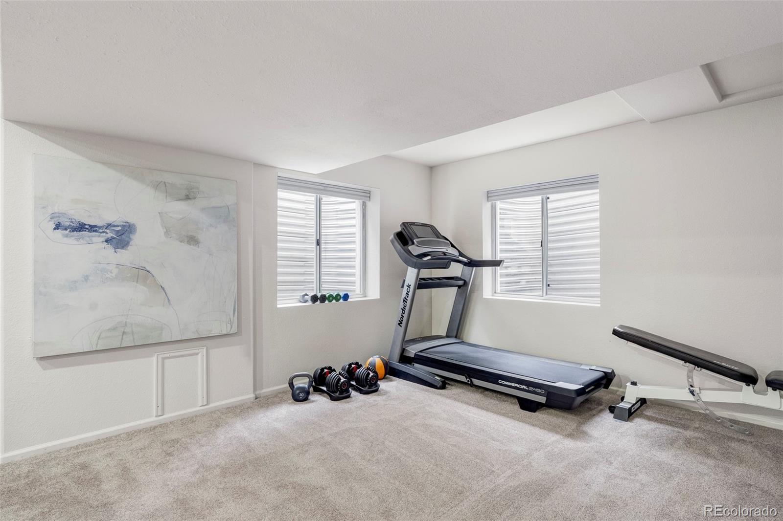 Home gym with a treadmill, free weights, and a weight bench beneath two windows.