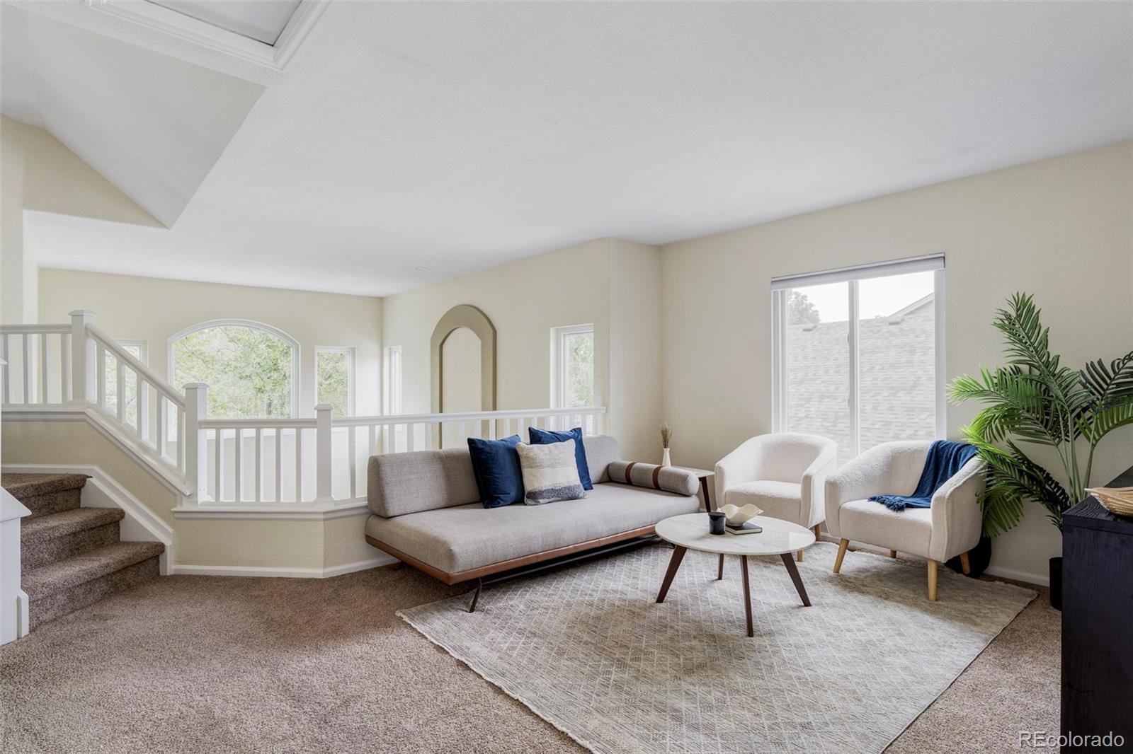 Staged living room with a gray sofa, white armchairs, and staircase overlooking the seating area.