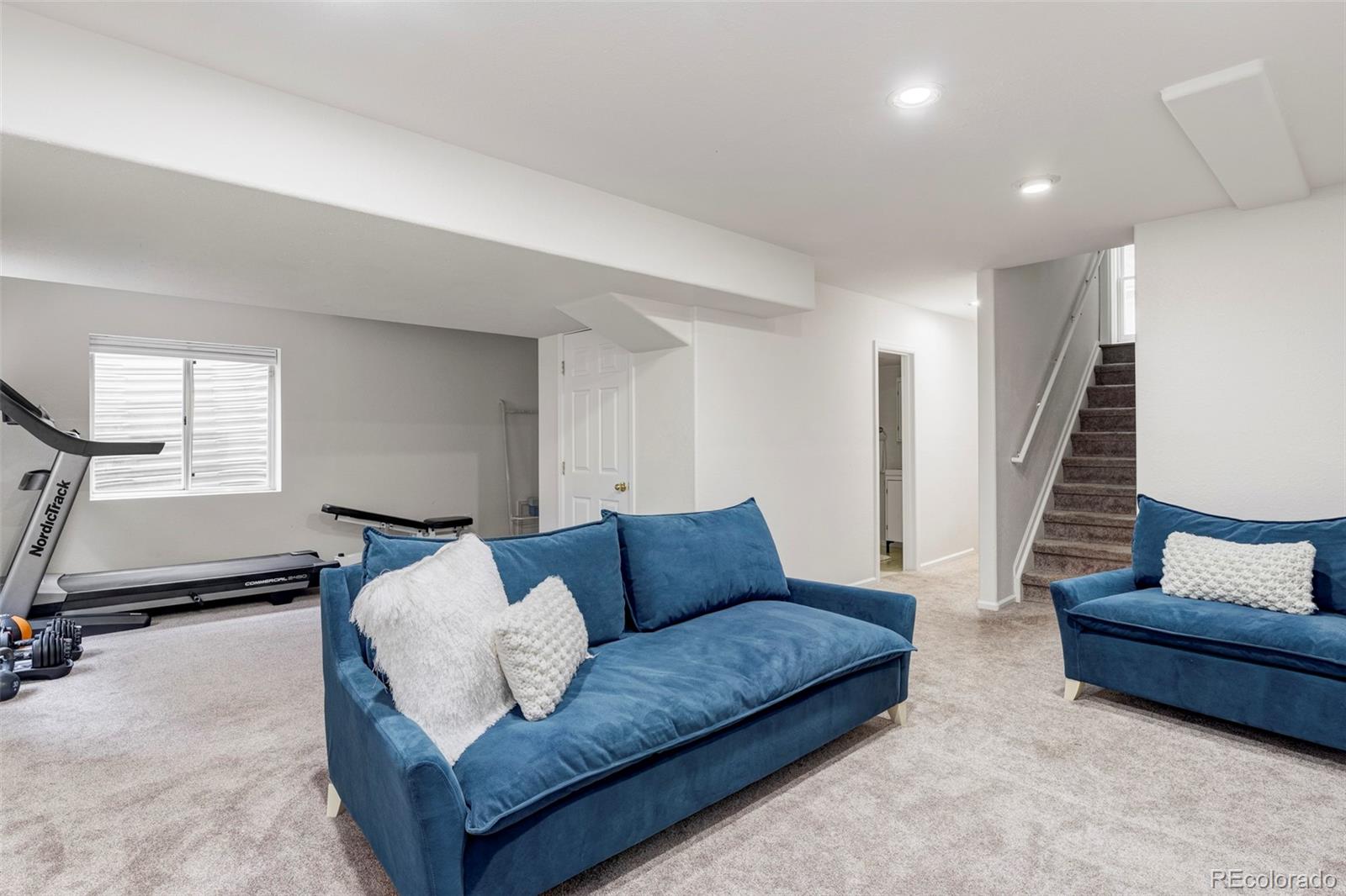 Basement living area with blue sofas, exercise equipment, and carpeted stairs.